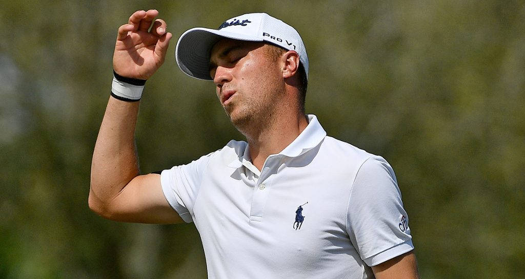 沮喪的 Justin Thomas 壁紙 — 一位專注的高爾夫球手，身穿白色Polo衫和帽子，站在翠綠的球場上，用手遮擋刺眼的陽光。