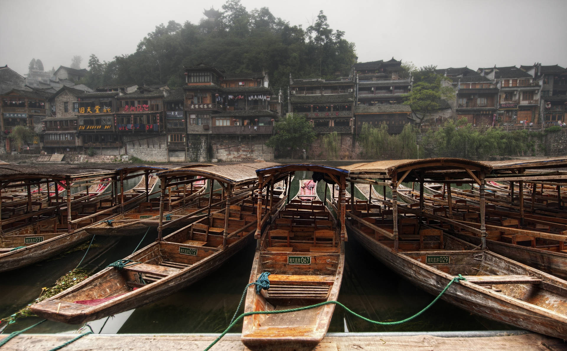 木船停泊在霧氣瀰漫的河邊，山坡上有傳統的中國建築。