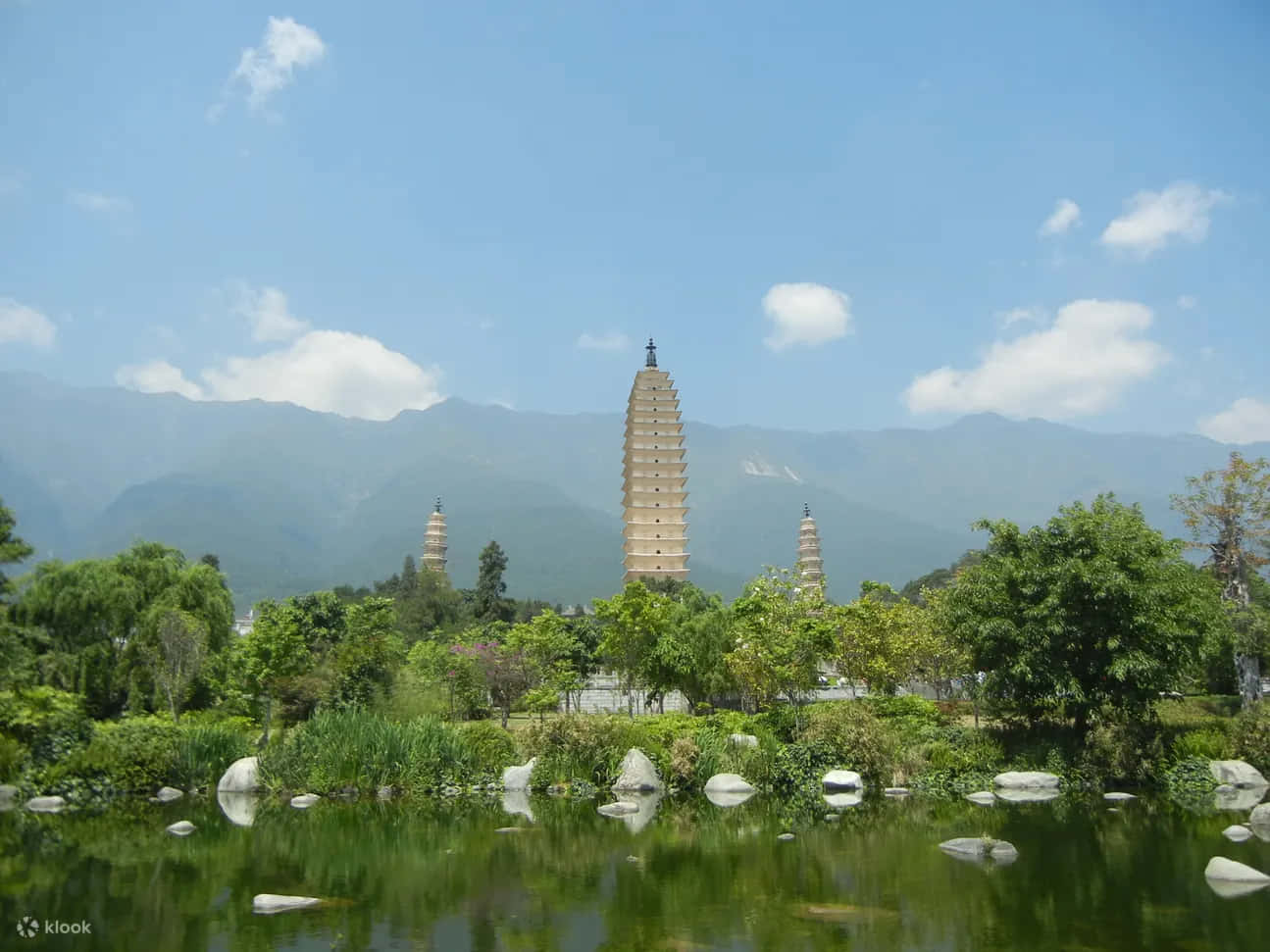 河畔岩石三塔 壁紙 — 三座古塔聳立在茂密的綠植和寧靜的池塘之上，池塘倒映著清澈的藍天和遠山。