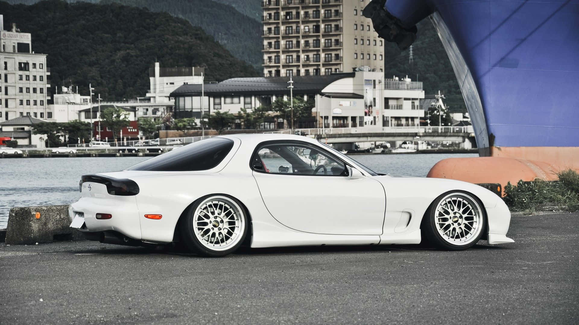 白色萬事得RX-7跑車停泊在水邊，背景是城市建築物。