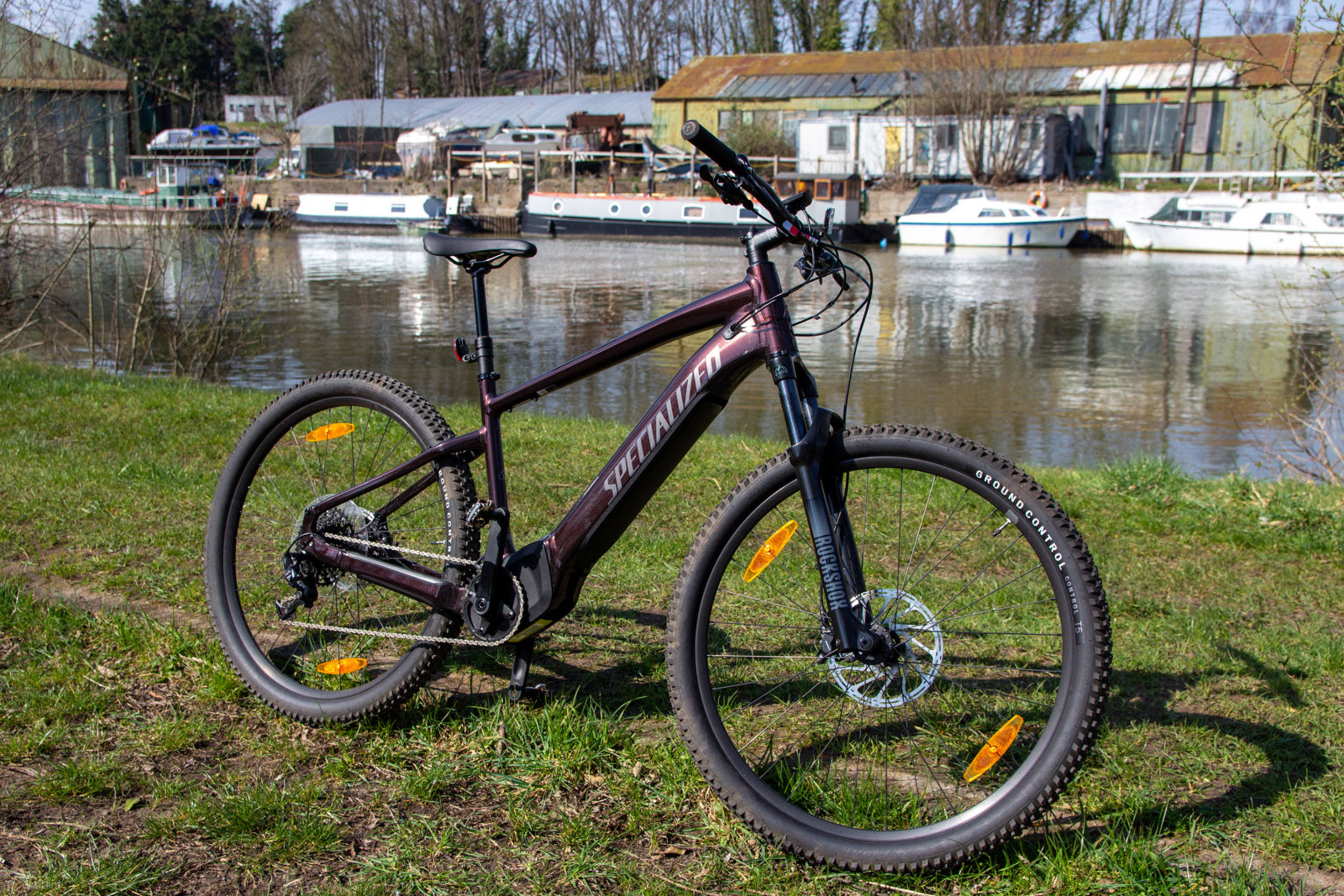 河畔的 Specialized 自行車 壁紙 — 一輛栗色電動山地單車停放在草地上，旁邊是一條平靜的河流，背景中有繫泊的船隻和質樸的建築物。