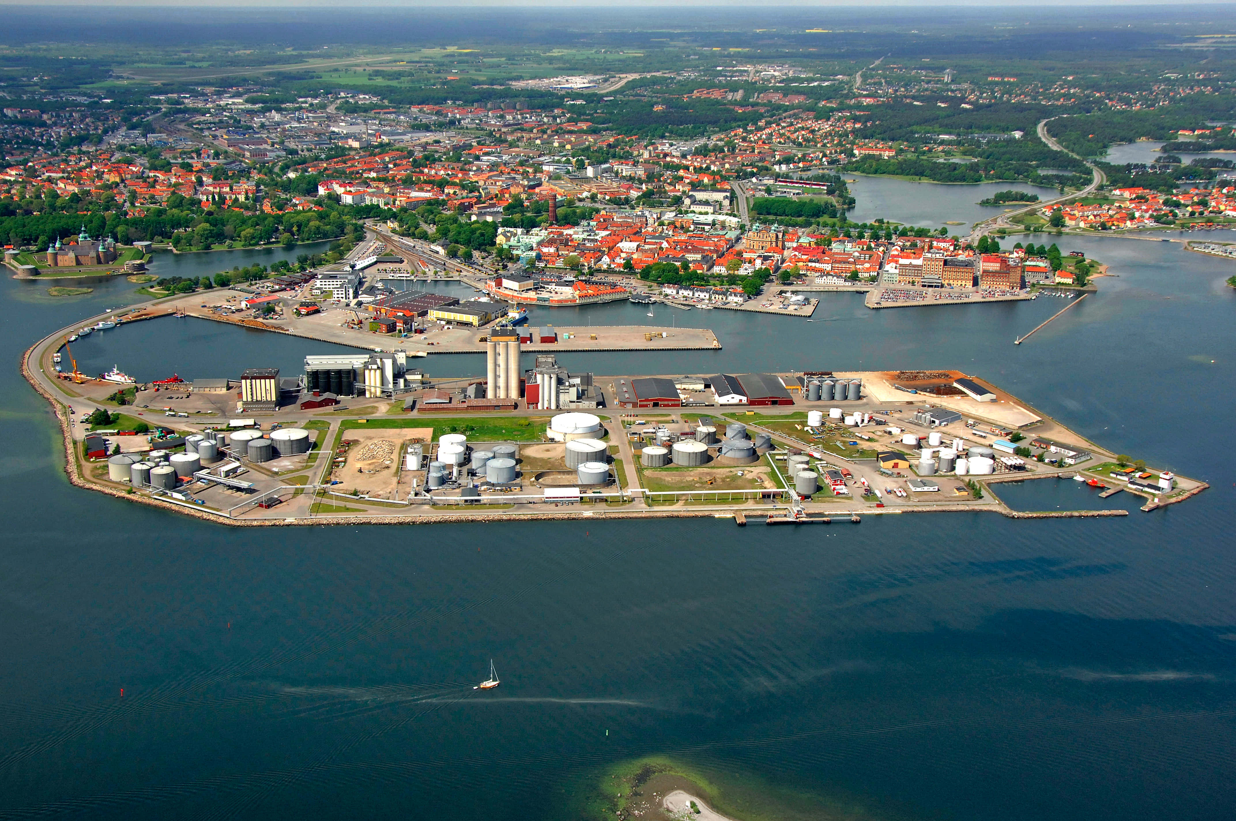 Kalmar Sweden 桌布 壁紙 — 沿海城市的鳥瞰圖，有紅頂建築和工業港口，設有大型銀色儲罐和帆船在平靜的藍色水面上。