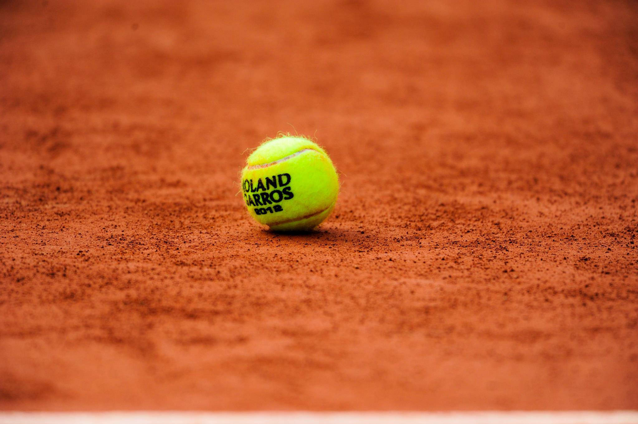 法國公開賽網球球聚焦特寫 壁紙 — 一個鮮黃色嘅網球，上面有 Roland Garros 2012 字樣，放喺有紋理嘅紅土球場表面上。