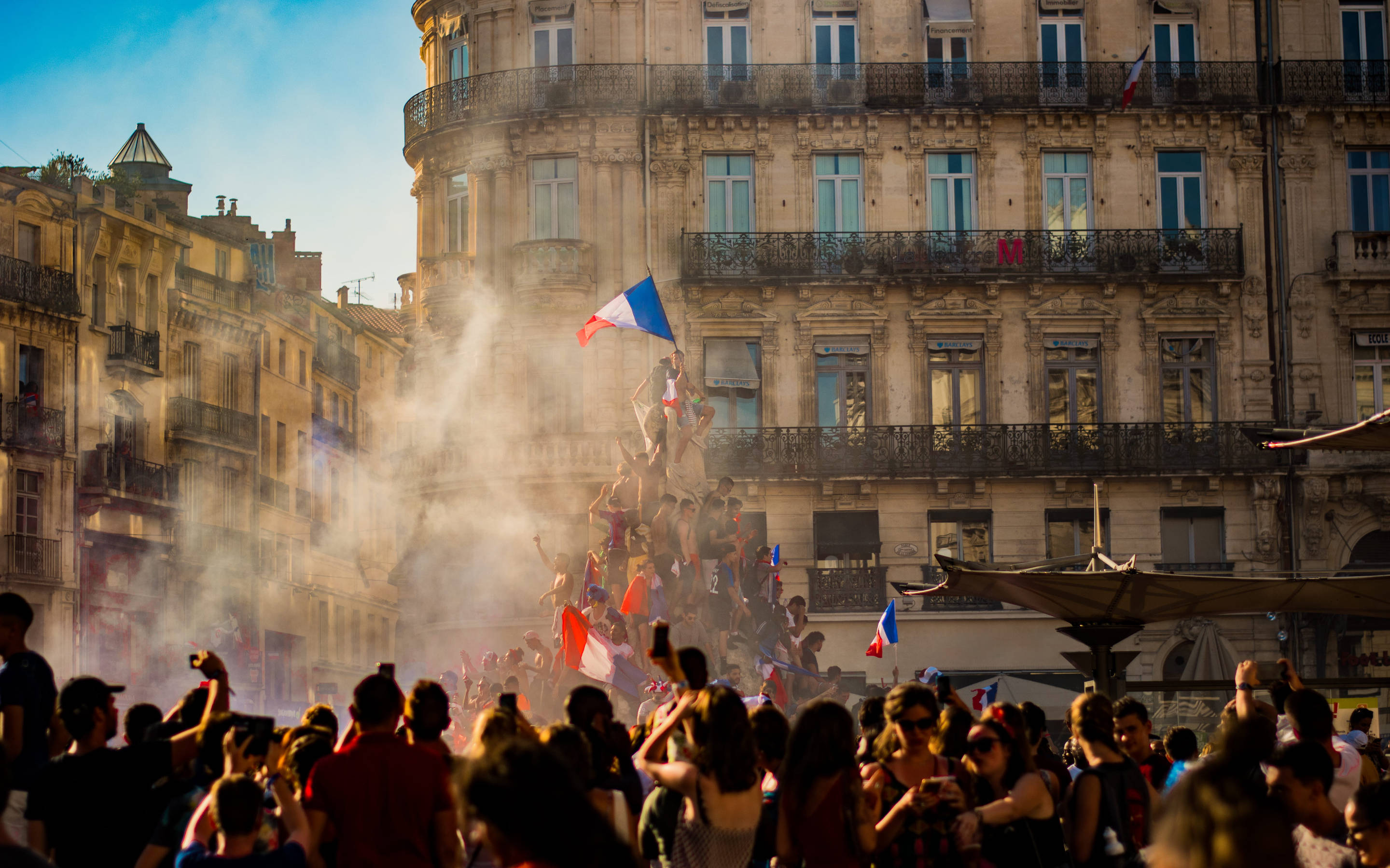 法國國旗肖像 壁紙 — 在陽光明媚的歐洲廣場上，歡呼的人群在歷史建築和煙霧中揮舞法國國旗慶祝。