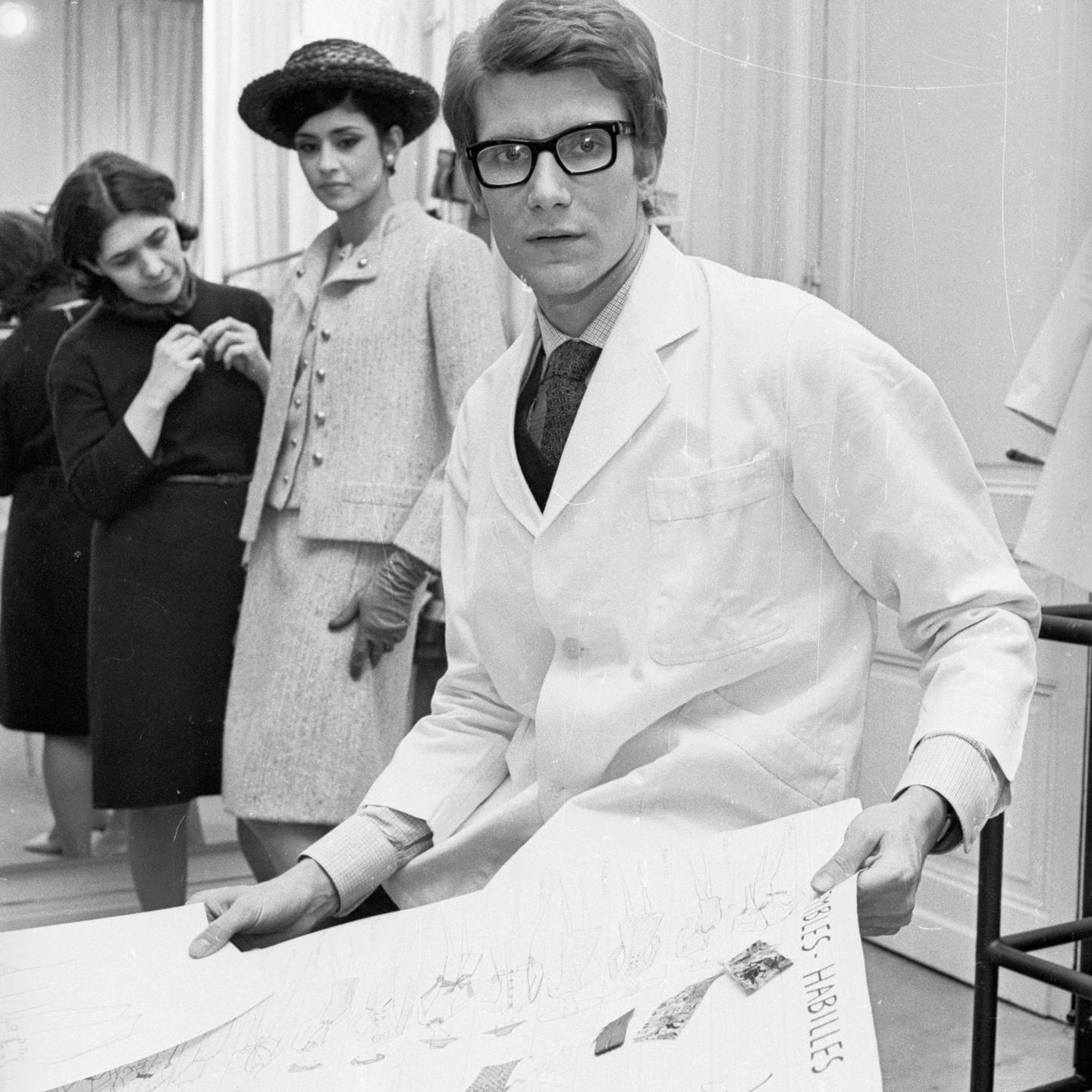 法國時尚設計師Yves Saint Laurent 壁紙 — 一名身穿白色實驗袍、戴著厚眼鏡的男子手持時裝草圖，兩名女子在復古工作室環境中觀察。