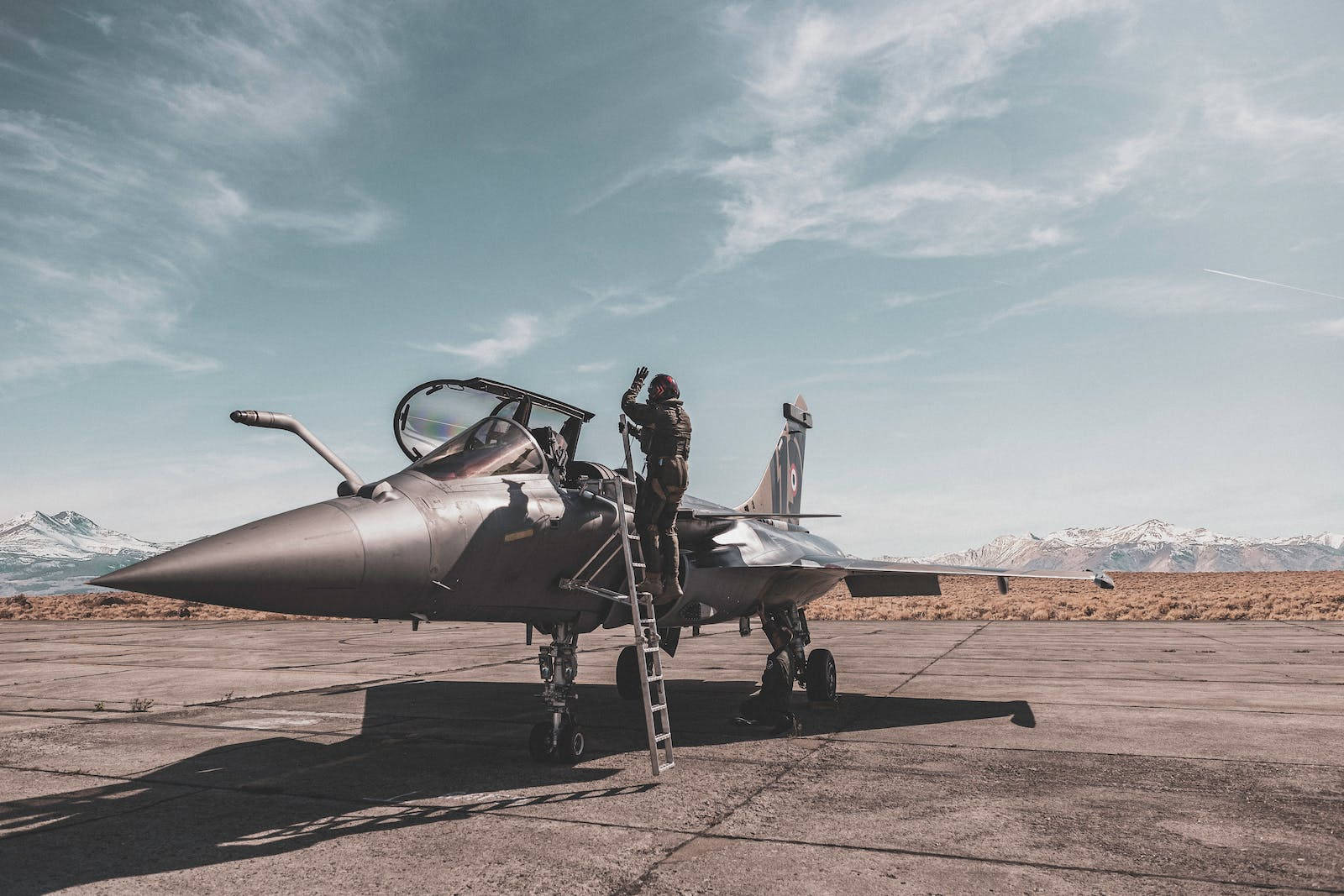 法國空軍戰鬥機 壁紙 — 一名戰鬥機飛行員爬上梯子，進入一架停在停機坪上的流線型噴射機，背景是積雪的山脈，天空部分多雲。