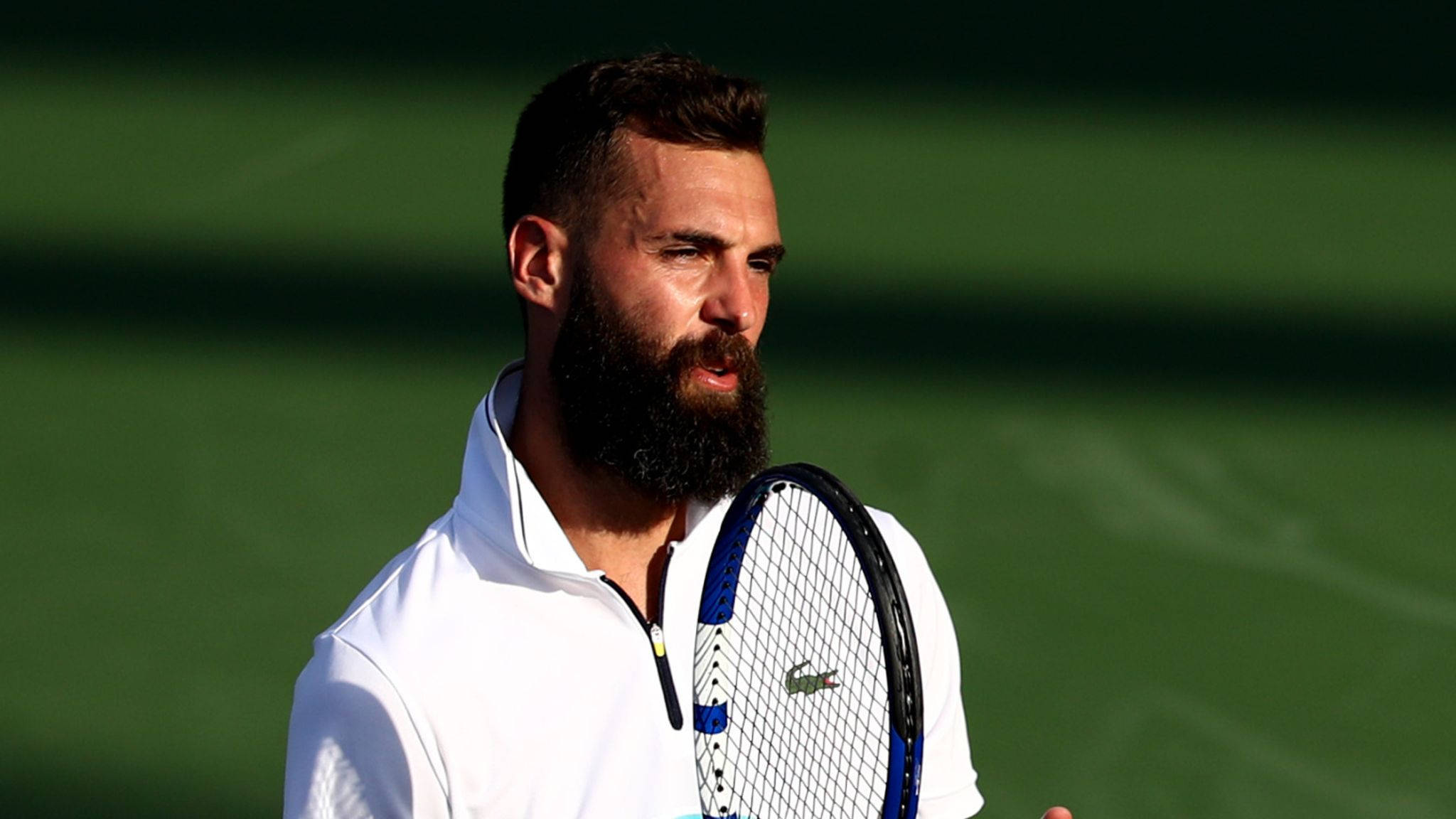 法國網球明星 Benoit Paire 場上揮拍 壁紙 — 一位留鬍鬚的男性網球選手穿著白色襯衫，在模糊的綠色球場背景下握著藍色球拍。