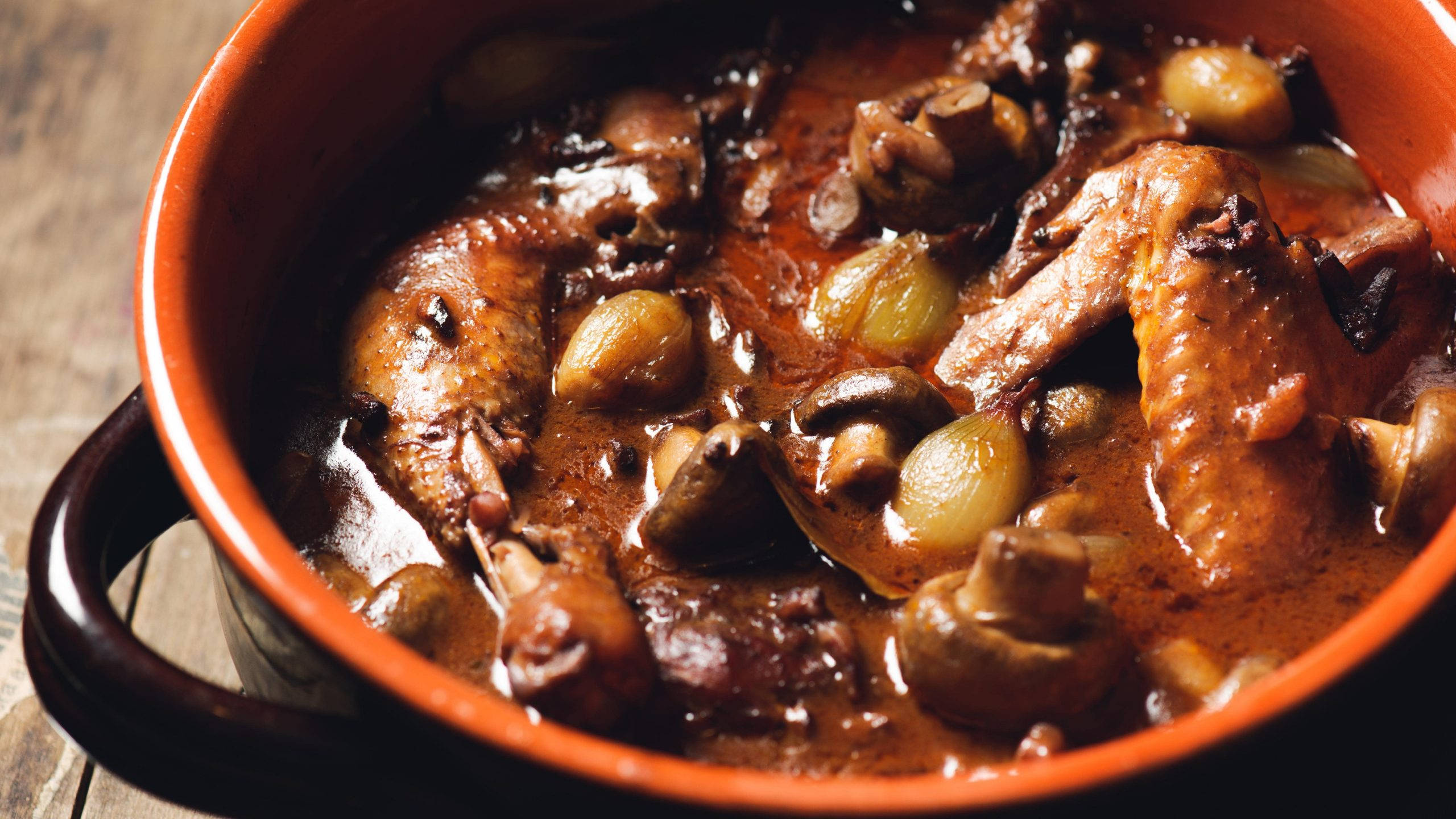 法式雞肉燉煮Coq Au Vin 壁紙 — 豐富的棕色燉菜，包含雞肉、蘑菇和洋蔥，在木質表面上的質樸橙色陶瓷鍋中慢燉。