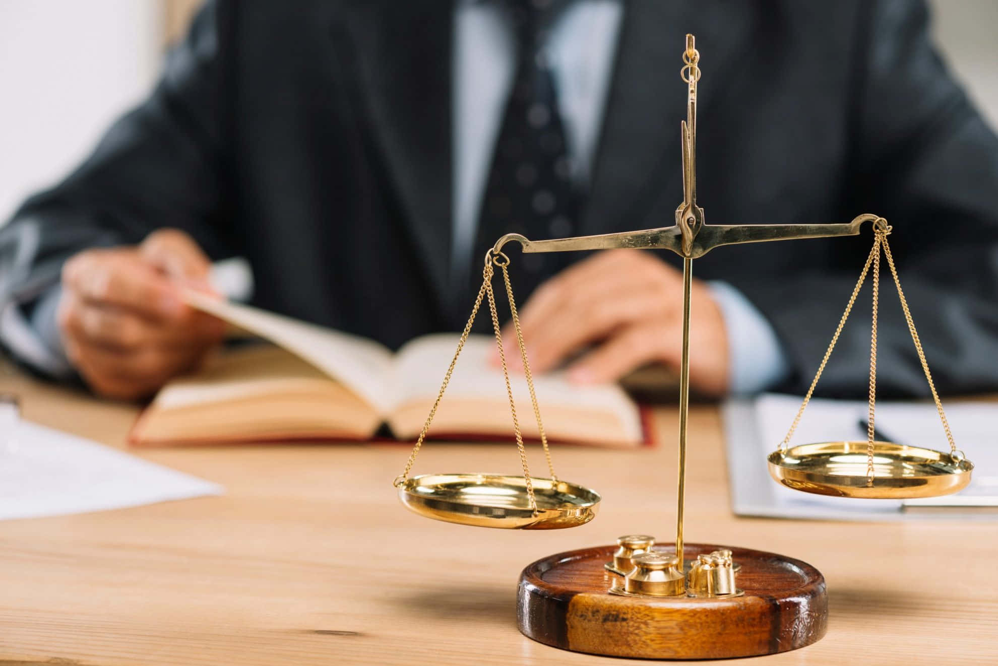 法律專業中，聰明工作勝過苦幹 壁紙 — 一個金色的正義天秤顯眼地放在木桌上，背後有一位穿著西裝的模糊律師正在寫書。