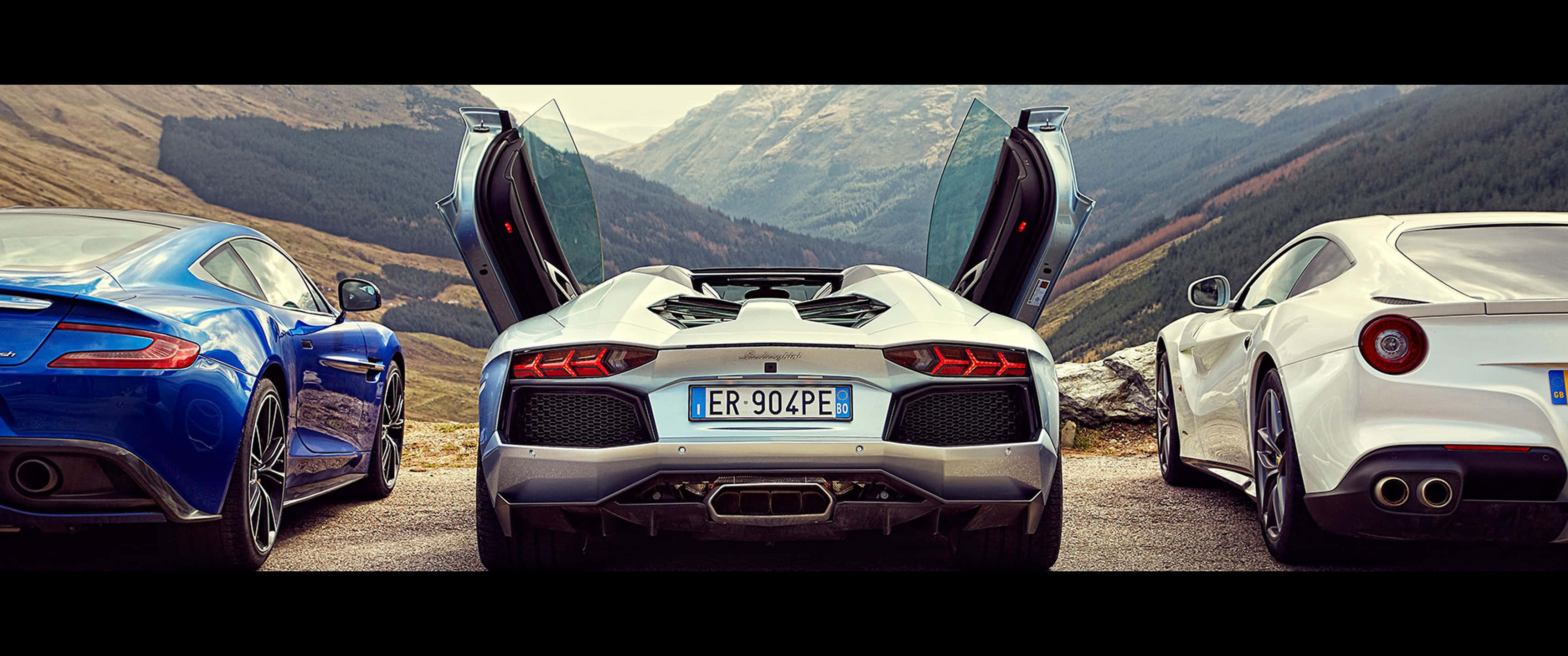 法拉利F12、蘭博基尼Aventador與阿斯顿·马丁Vanquish山景背景（3440x1440p） 壁紙 — 三輛豪華跑車打開車門停泊在山路上，背景是崎嶇的阿爾卑斯山。