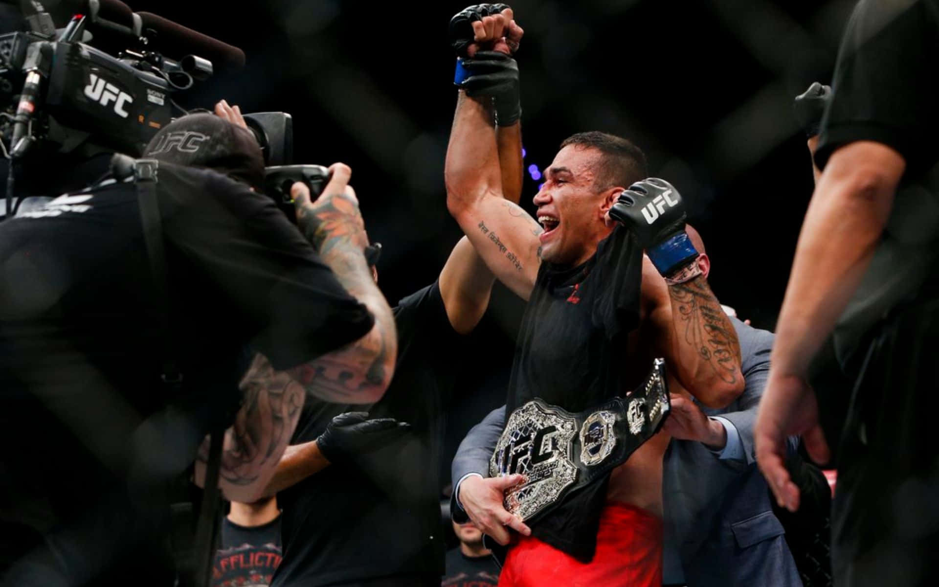 法比西奧·韋德姆慶祝UFC 188勝利 壁紙 — 一位勝利的UFC拳手在八角籠內被攝像機和工作人員包圍，高舉冠軍腰帶和拳頭慶祝勝利。