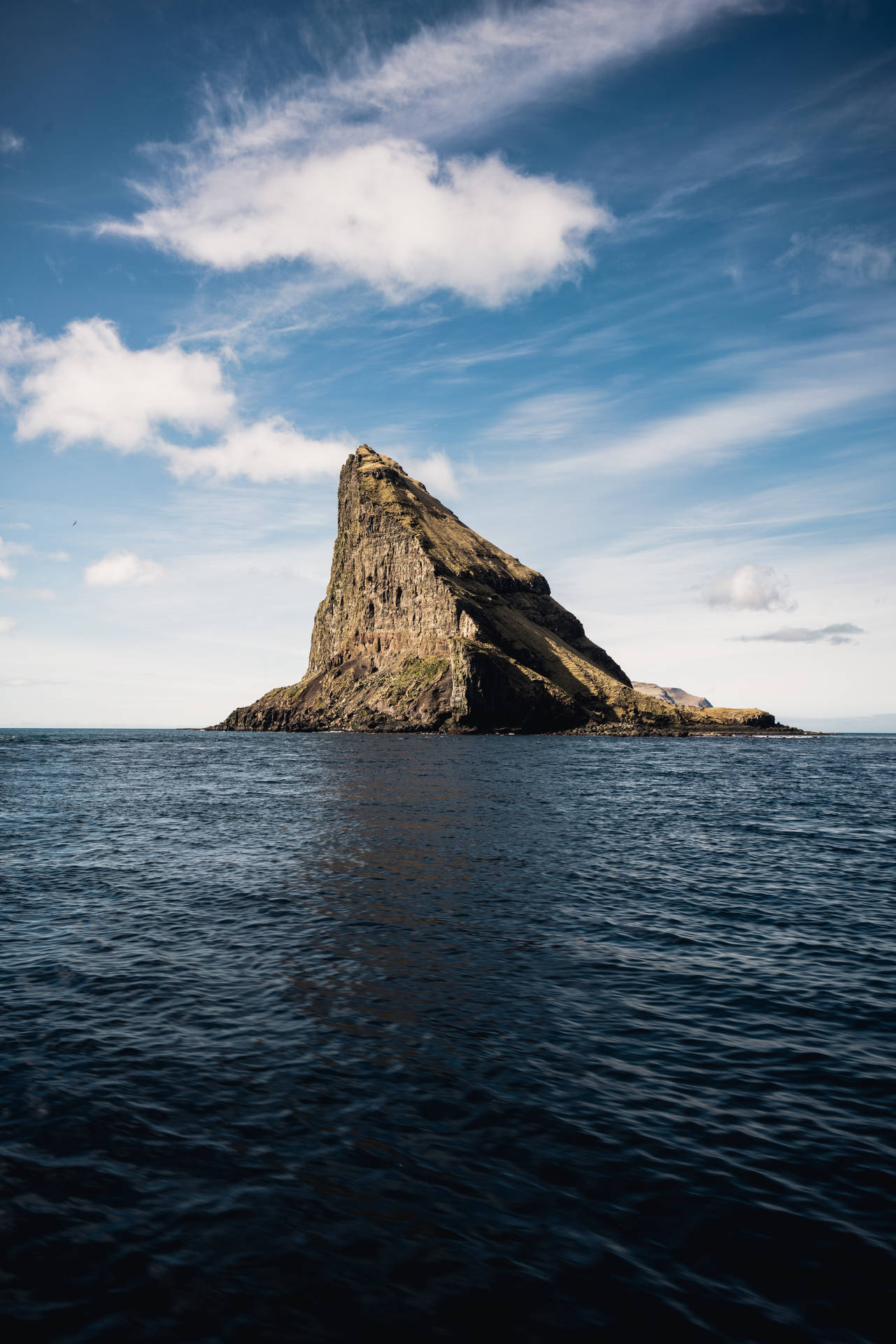 一座陡峭的岩石島嶼獨自矗立在開闊的海洋中，天空部分多雲。