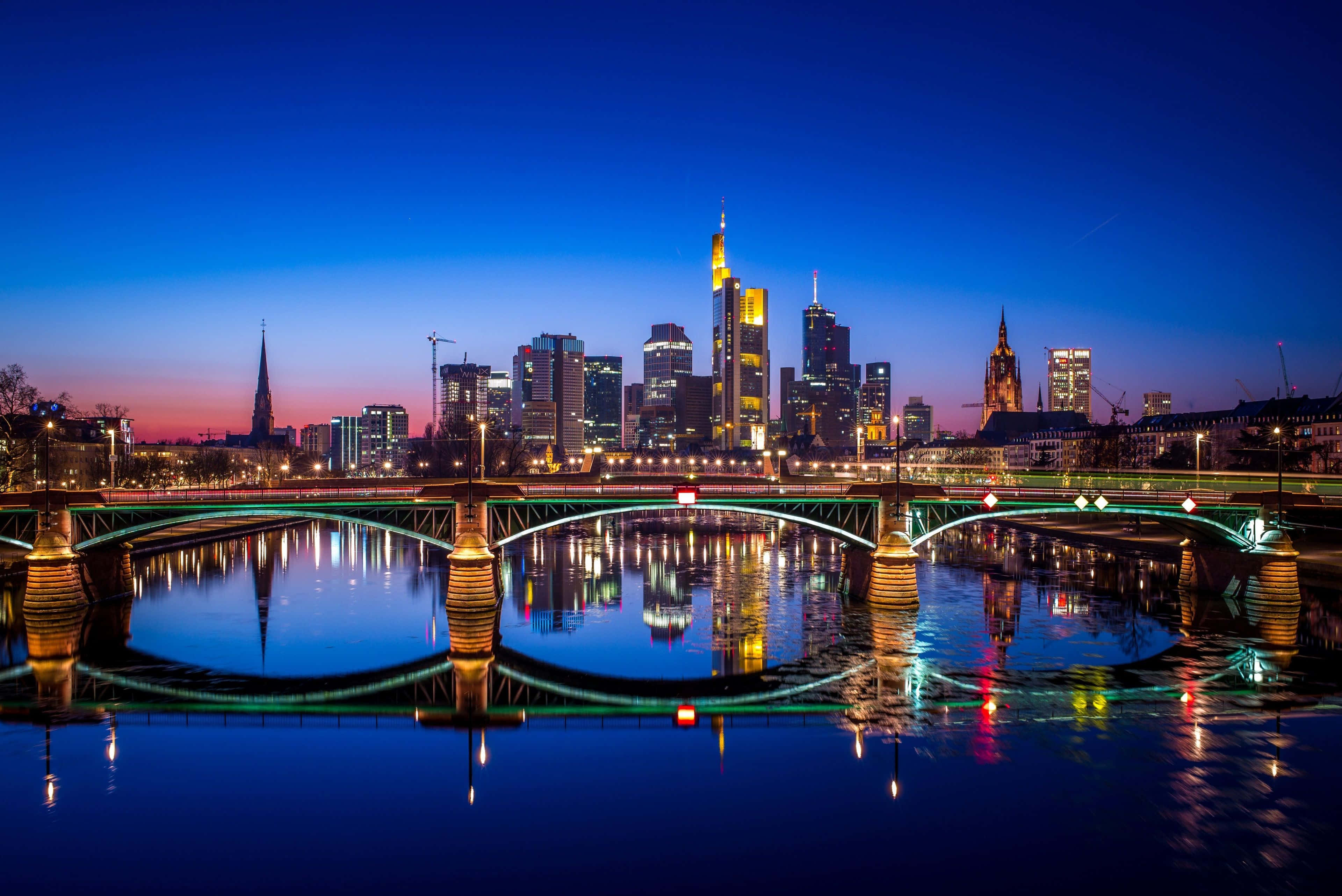 Germany 桌布 壁紙 — 法蘭克福令人驚嘆的黃昏城市景觀，燈火通明的摩天大樓和橋樑倒映在平靜的河水上。