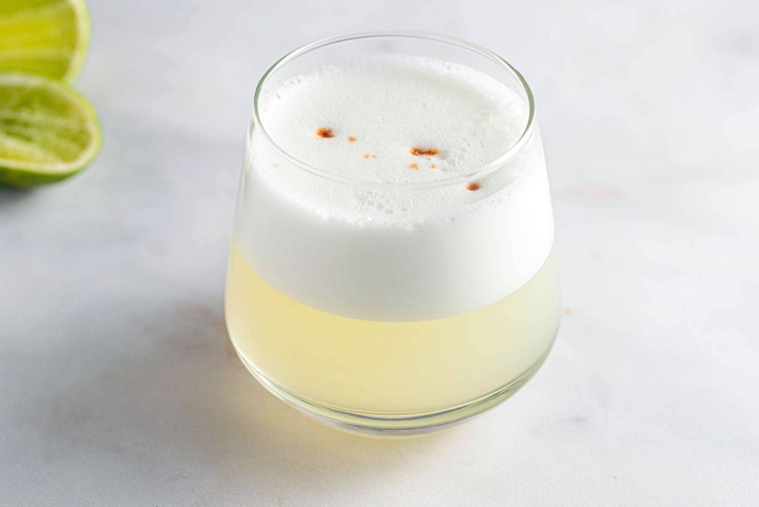泡沫酸味雞尾酒 壁紙 — 一杯泡沫狀的淡黃色雞尾酒，上面有橙色香料點和青檸角，放在大理石表面上。