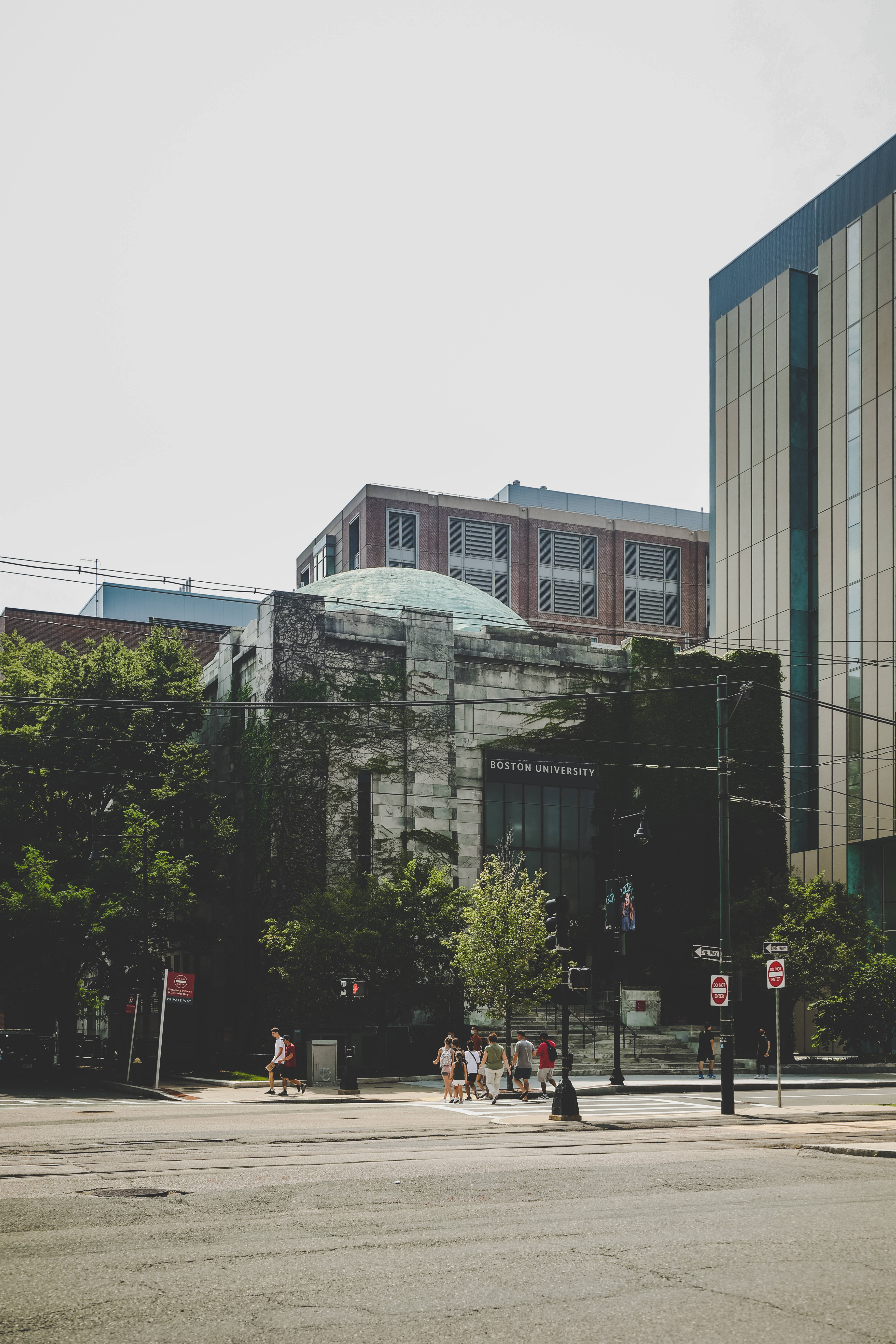 波士頓大學 壁紙 — 陽光明媚的城市街景，行人正在波士頓大學一棟被常春藤覆蓋的歷史建築附近過馬路。