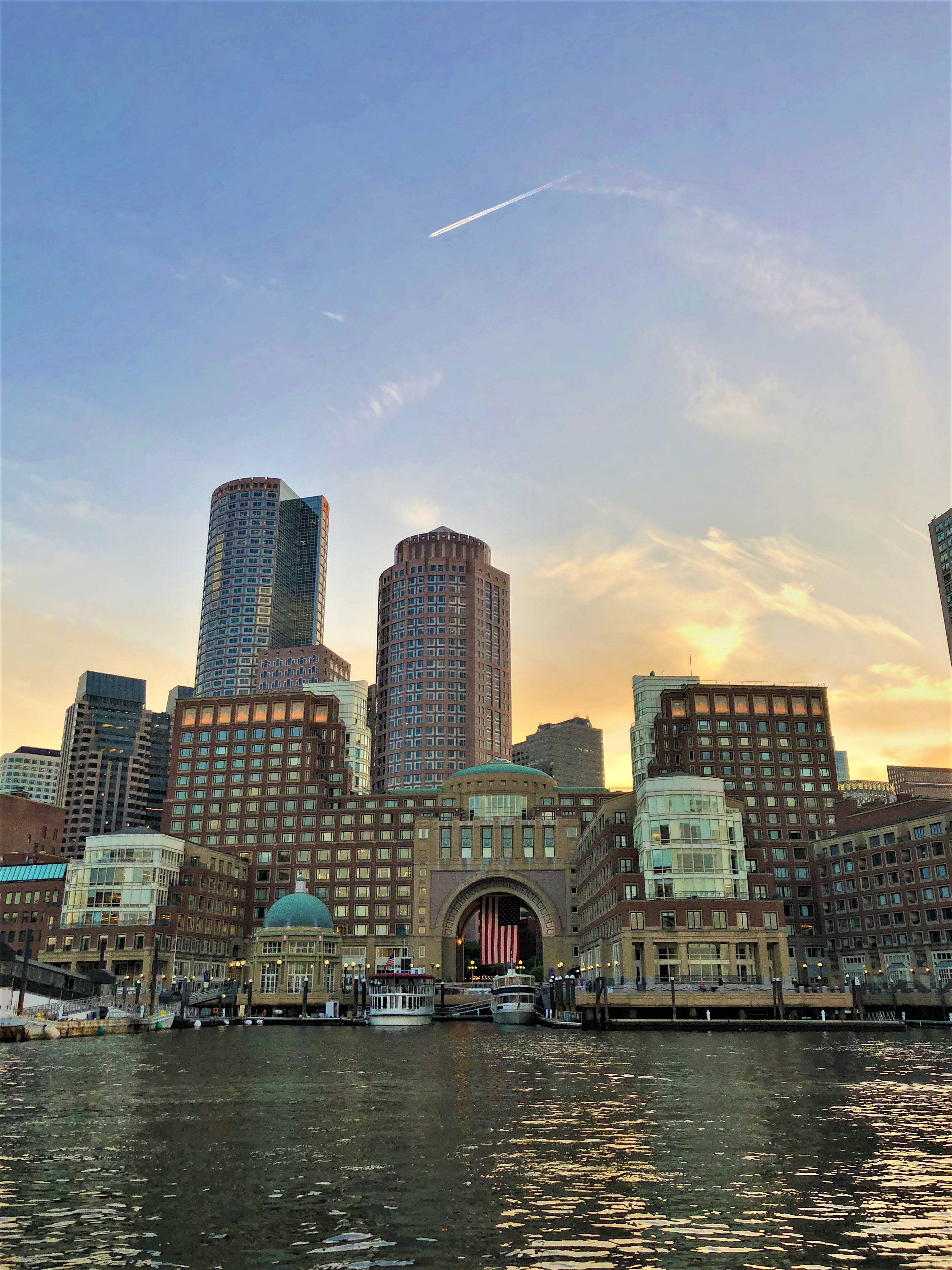 波士頓灣區建築 壁紙 — 夕陽將波士頓海濱天際線沐浴在金色光芒中，船隻在平靜的水面上休憩，天空劃過一道凝結尾跡。