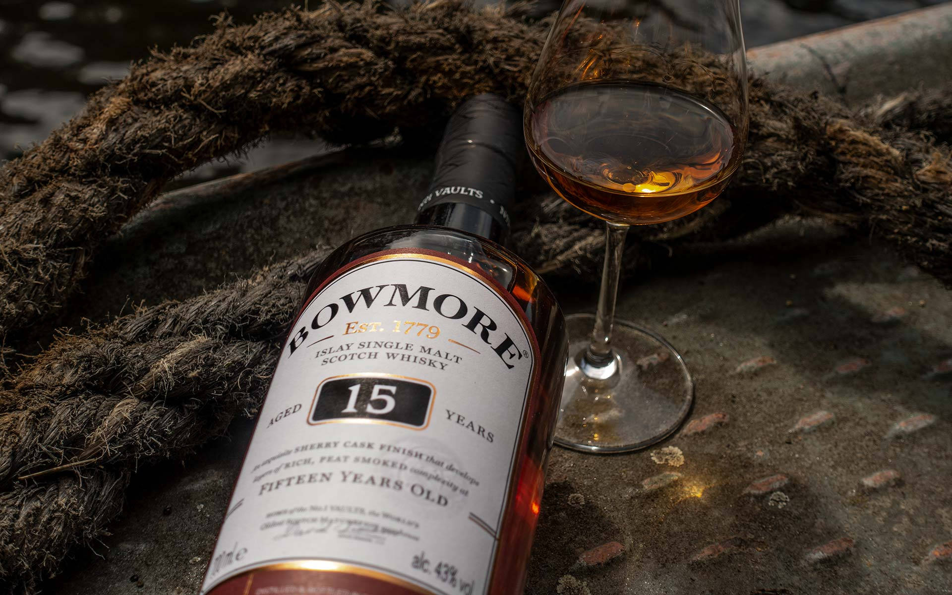 一瓶Bowmore 15年威士忌和一個玻璃杯放在粗繩旁邊的木頭上。