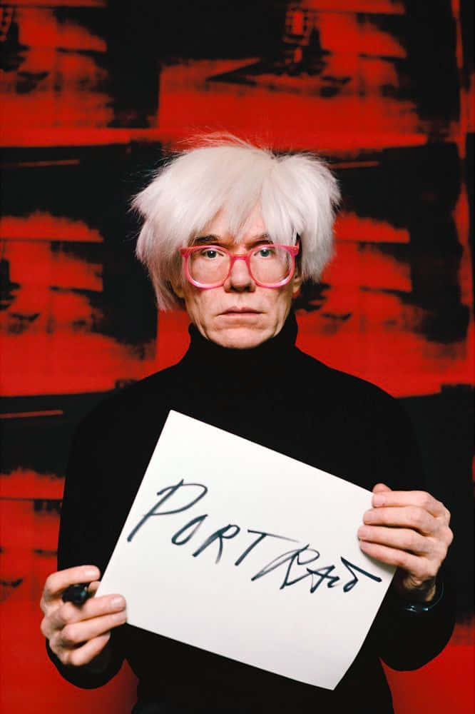 波普藝術偶像安迪·沃霍爾 壁紙 — Andy Warhol 白髮粉紅眼鏡，手持寫有「PORTRAIT」的標語牌，背景為大膽的紅黑抽象圖案。