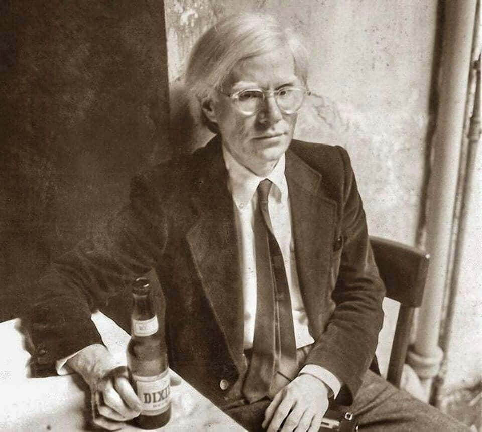 波普藝術先驅安迪·沃荷拍照 壁紙 — 一張安迪·華荷的黑白肖像，他坐在桌子旁，手持啤酒瓶，表情嚴肅。