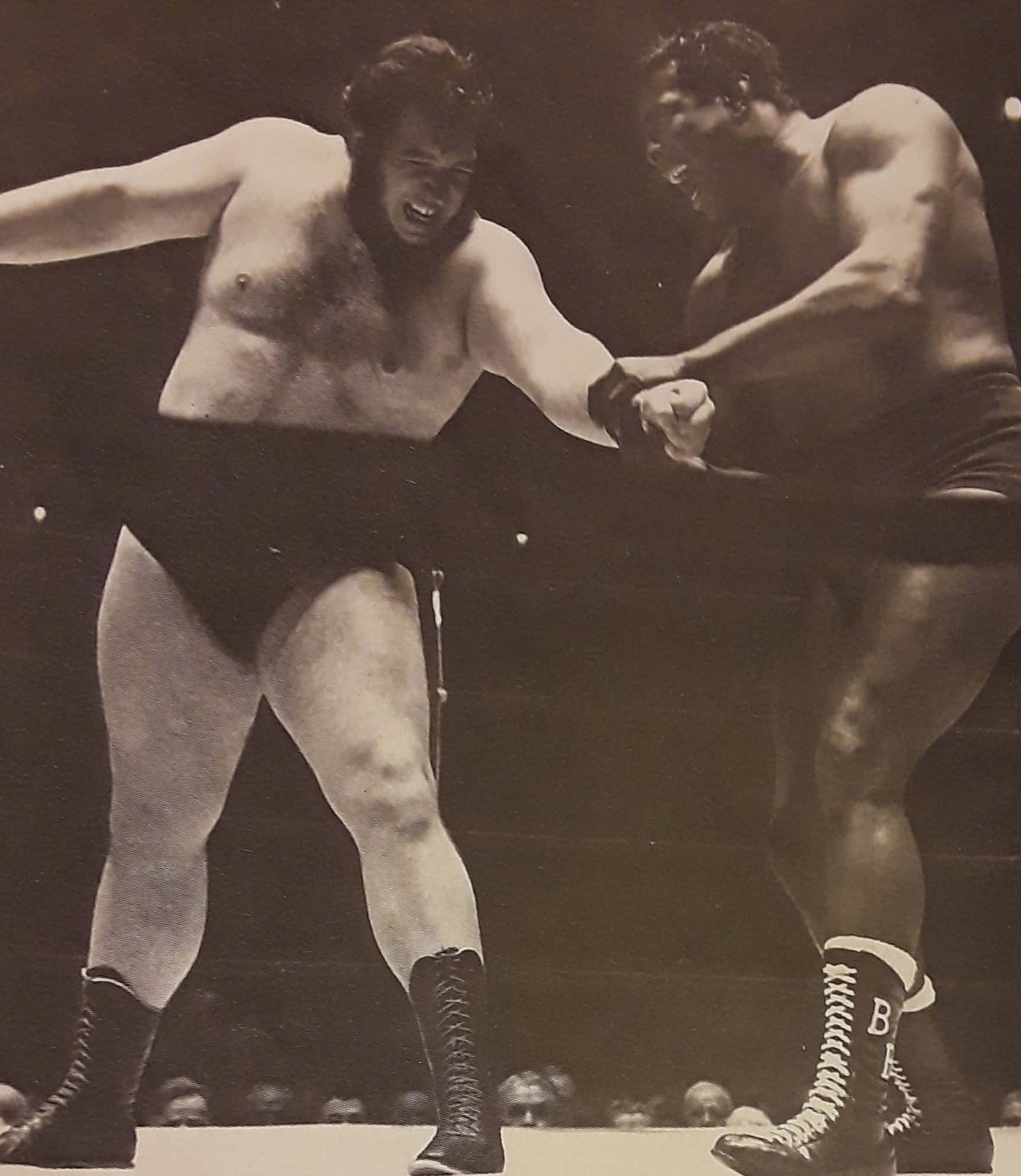 波波·巴西1963年對戰Gorilla Monsoon 壁紙 — 兩名肌肉發達的摔角手在燈光昏暗的擂台上激烈纏鬥，背景觀眾模糊不清。