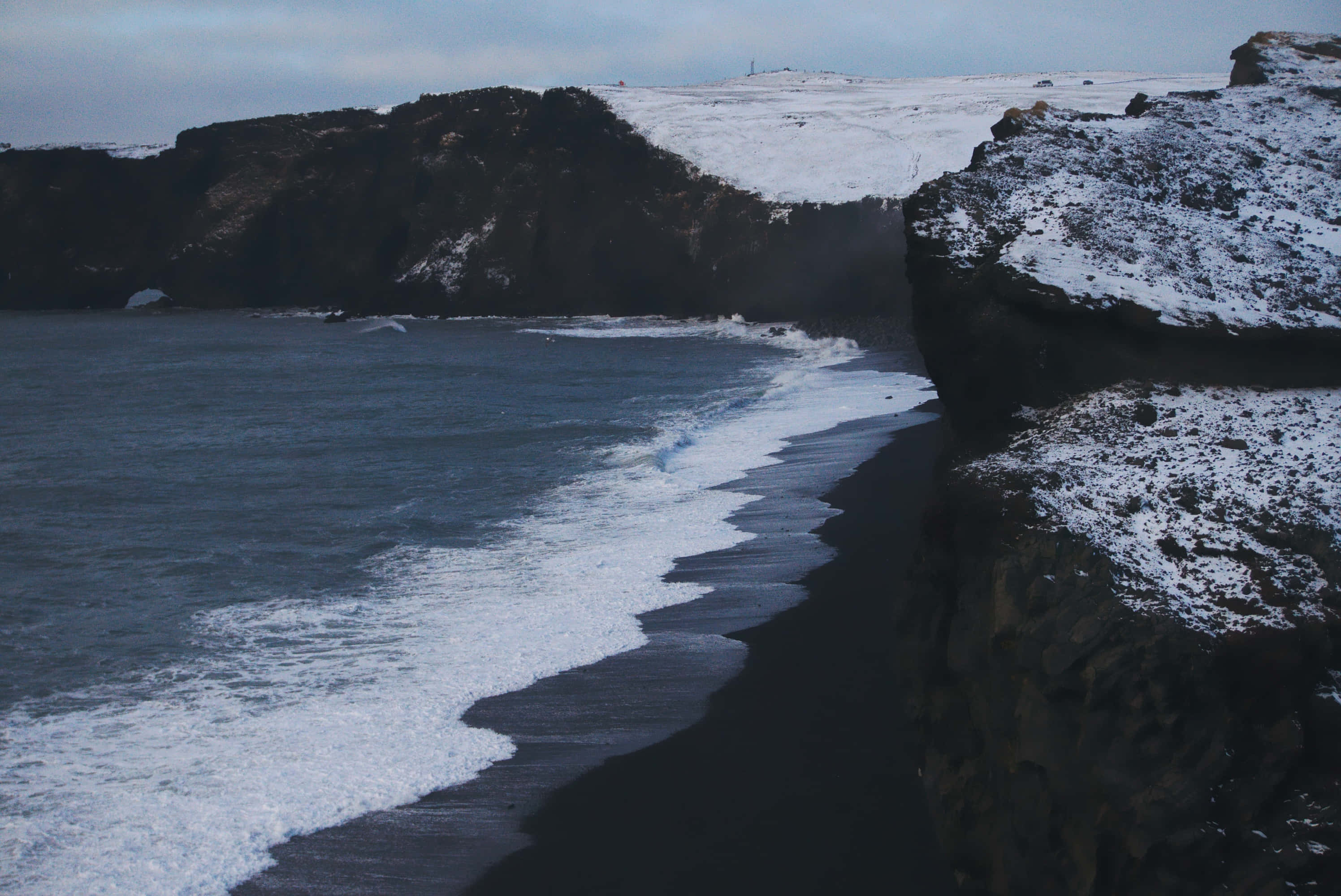 Black Sand Beach 桌布 壁紙 — 波浪沖上被雪覆蓋懸崖環繞的黑色火山沙灘，天空多雲。