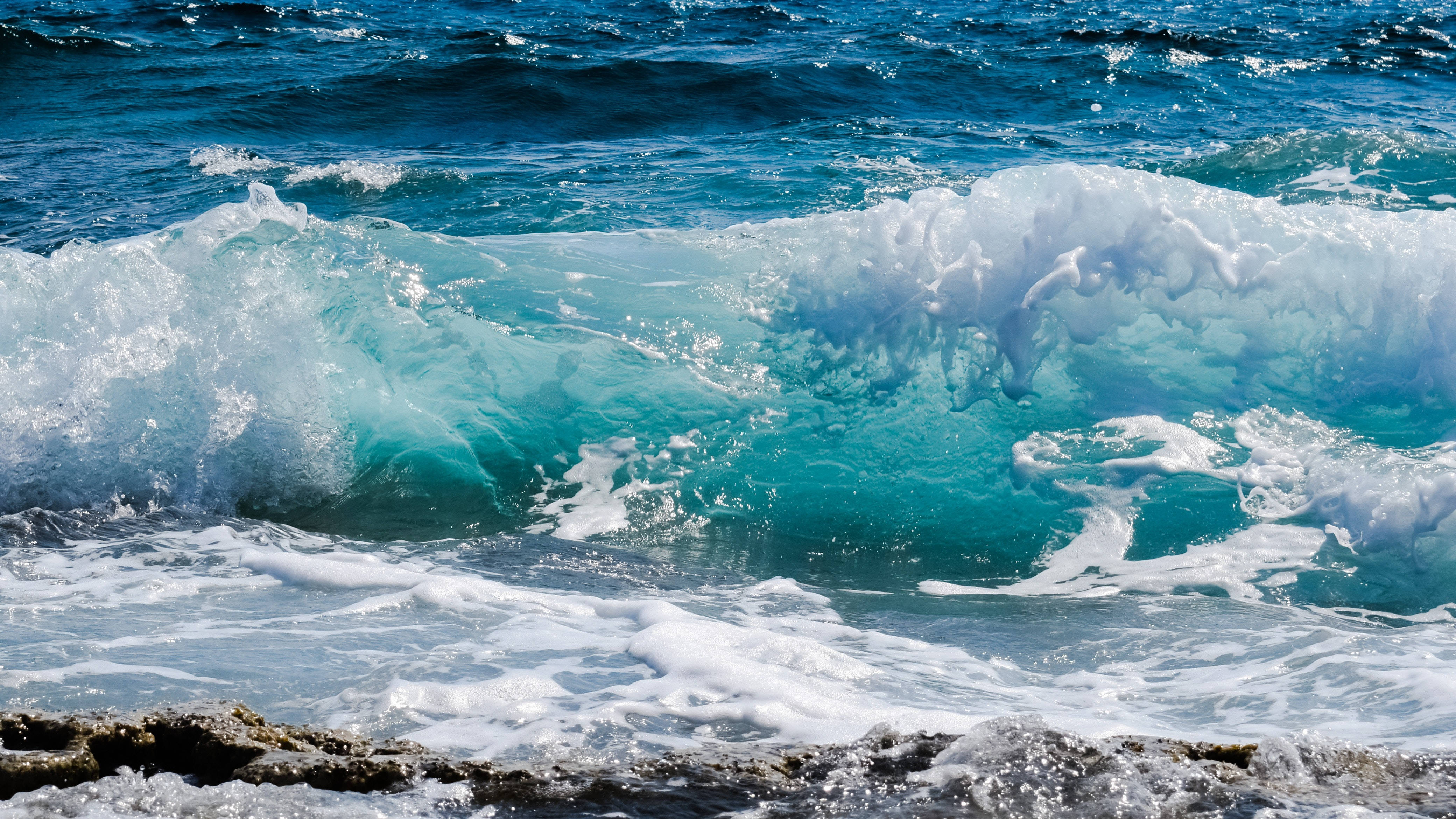 波濤洶湧的海景背景 壁紙 — 一道強勁的碧綠色海浪撞擊在深色岩石上，白色泡沫在明亮的日光中飛濺。