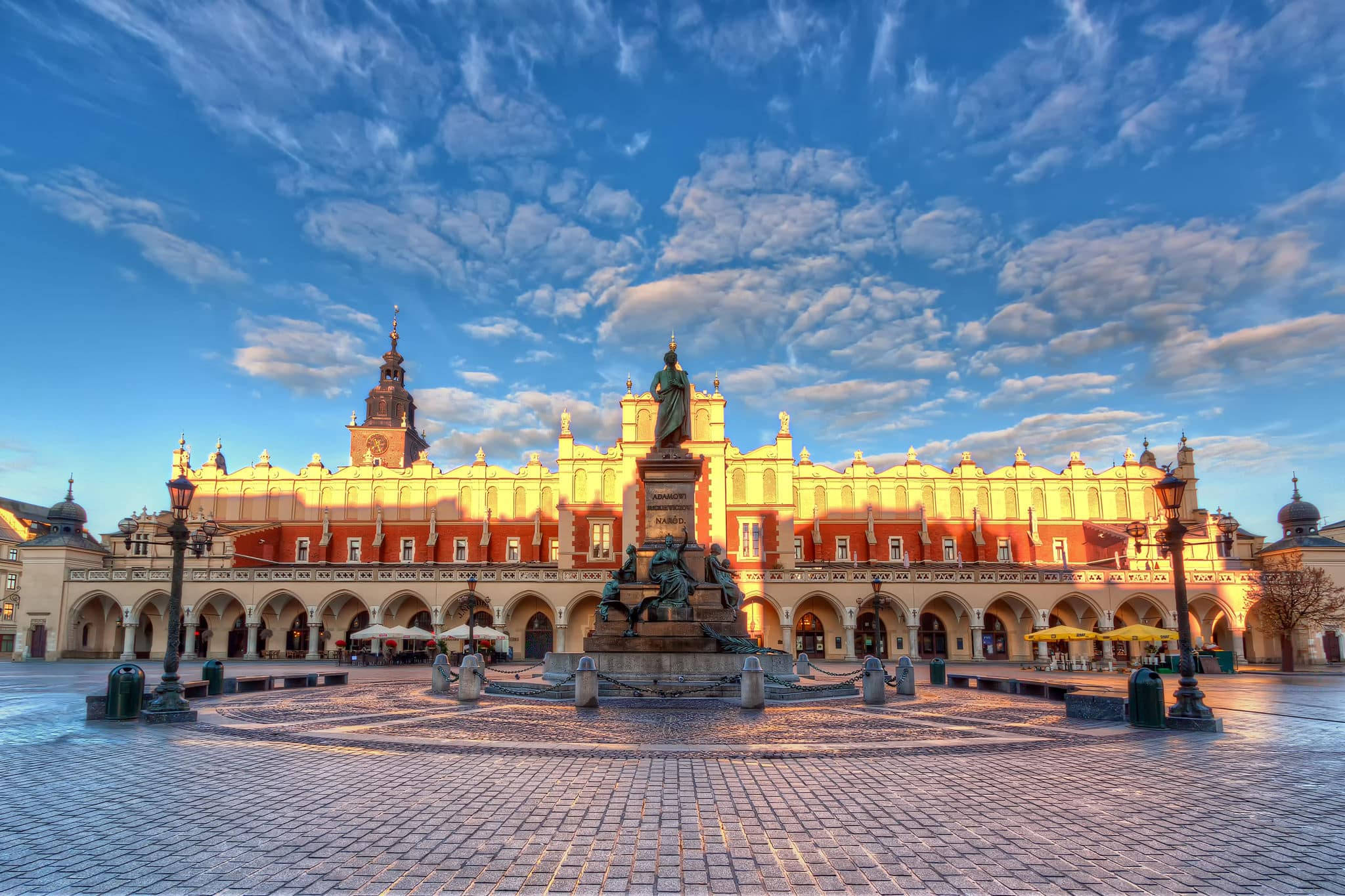 波蘭克拉科夫主廣場 Rynek Główny 壁紙 — 一個宏偉的歷史廣場沐浴在金色的日出光芒中，中央有一座雕像和華麗的建築，在充滿活力的藍天下飄著縷縷雲彩。