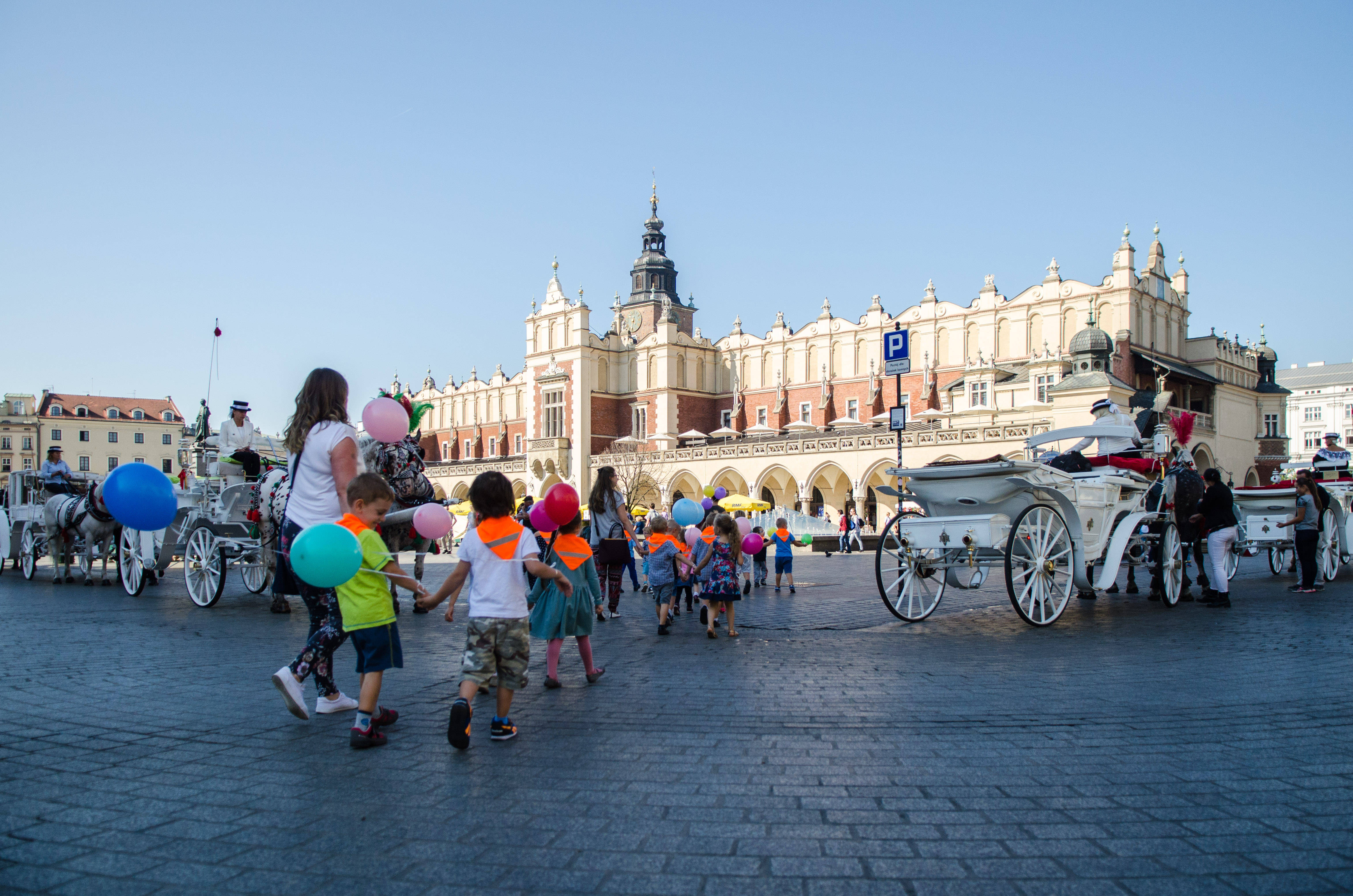 波蘭克拉科夫布料市場 壁紙 — 孩子們手持彩色氣球，在晴朗的藍天下，漫步於鵝卵石廣場，周圍有華麗的歷史建築和馬車。