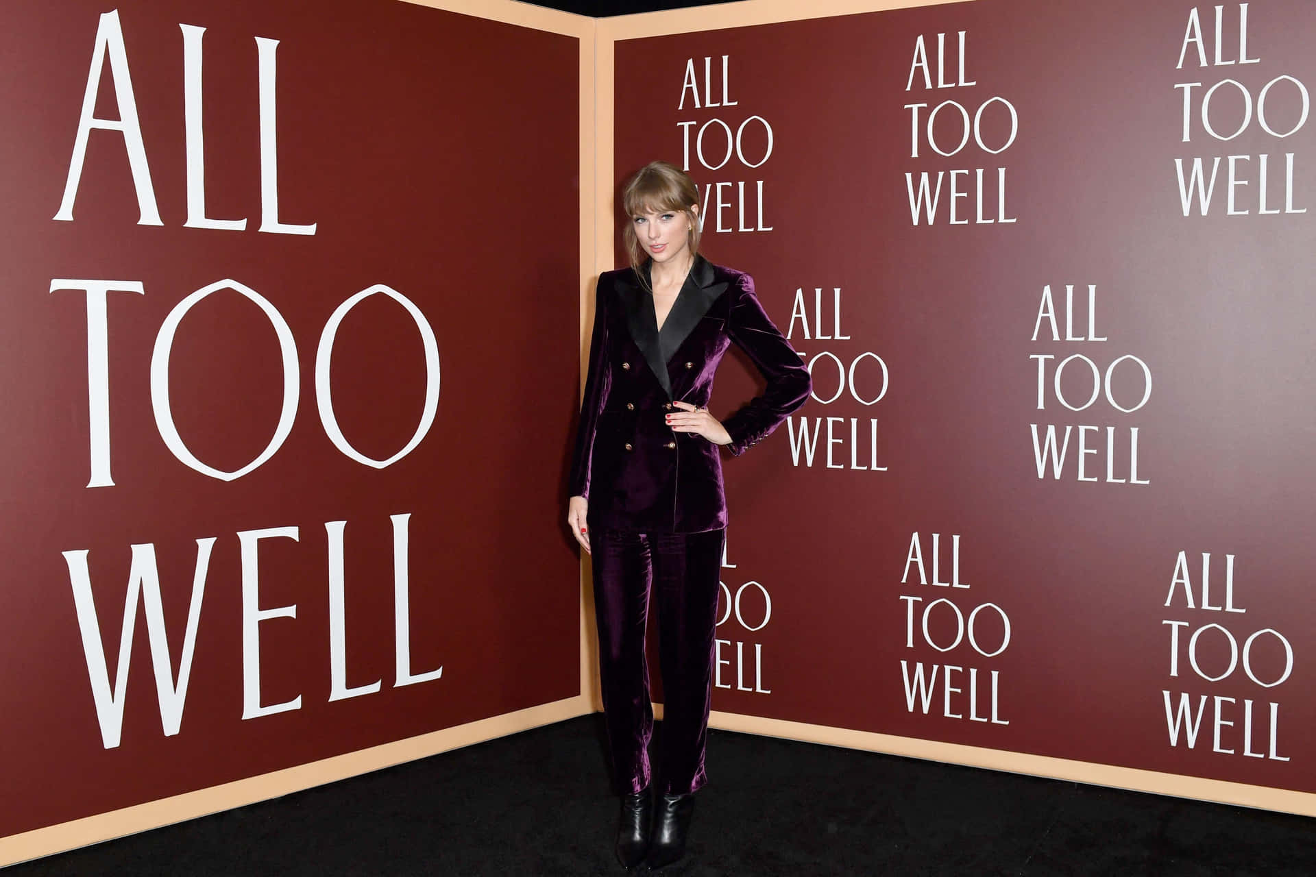 Taylor Swift 身穿紫色天鵝絨西裝，在一個有「ALL TOO WELL」文字牆的活動中擺姿勢。