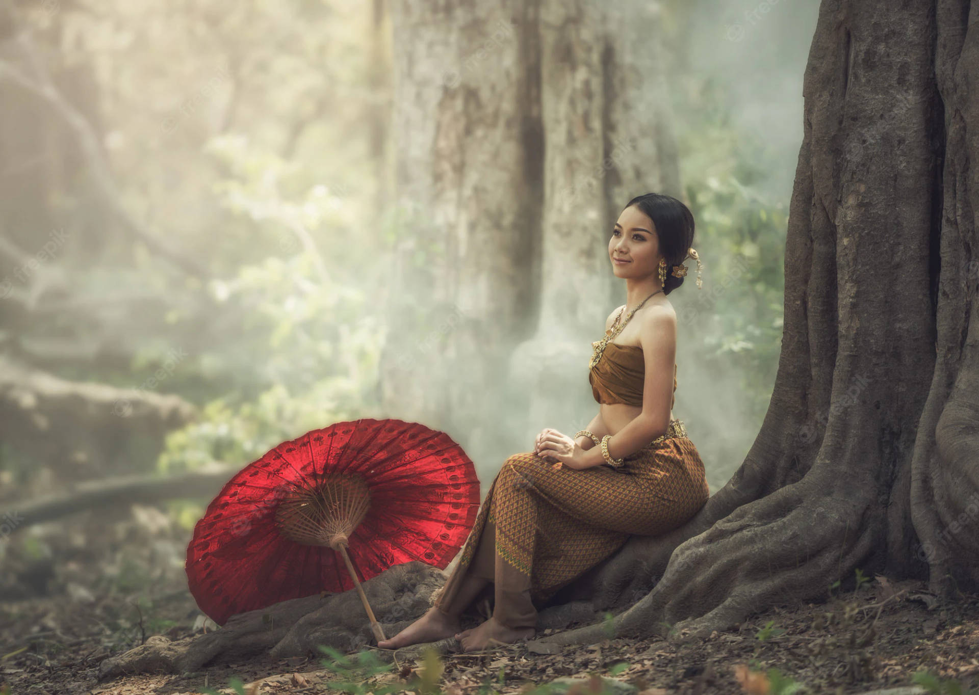 一位穿著傳統泰國裙的女性在迷霧森林中，拿著紅色陽傘坐在一棵大樹旁。
