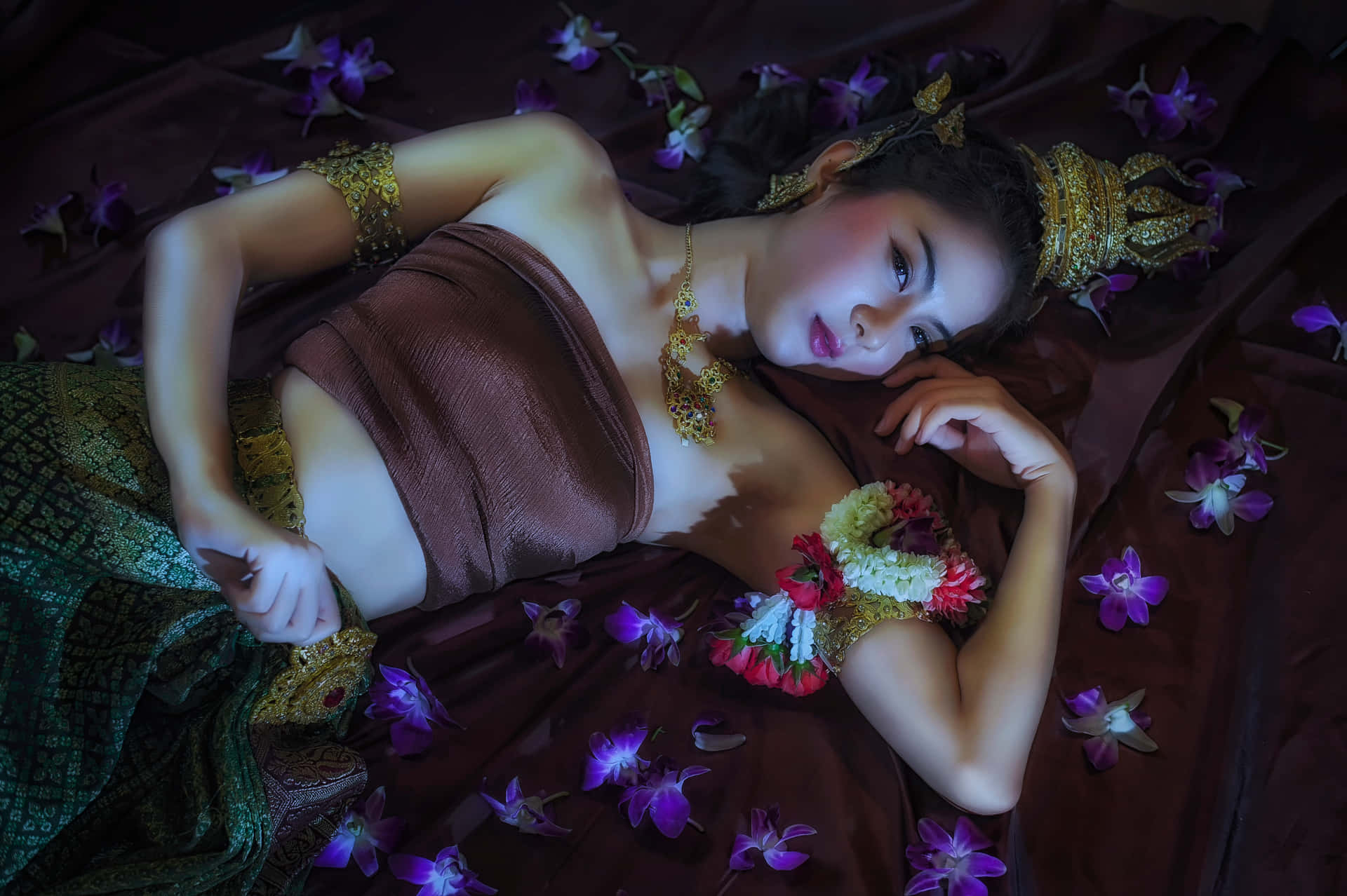 一位身穿傳統金色和棕色服飾的女性，在紫色蘭花床上休息。