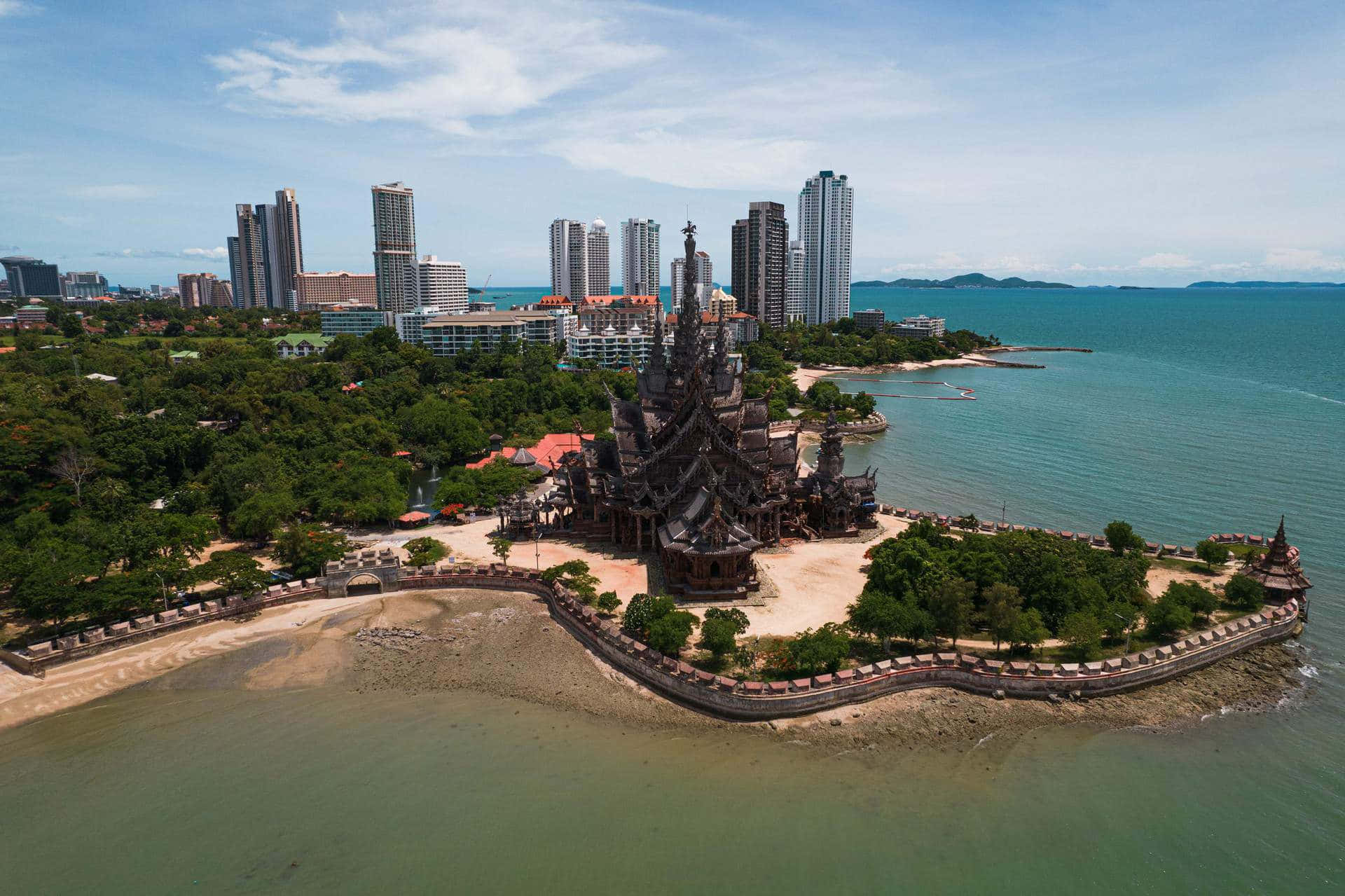 泰國海灘旁的真理聖殿 壁紙 — 海岸半島上深色木造真理寺廟的鳥瞰圖，背景有現代摩天大樓和碧綠海水。