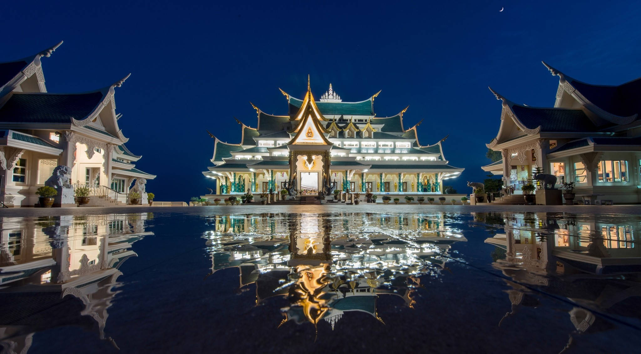 泰國烏東他尼瓦特帕富孔寺 壁紙 — 一個宏偉的泰國寺廟建築群在夜晚被照亮，完美地倒映在月光下的平靜水池中。