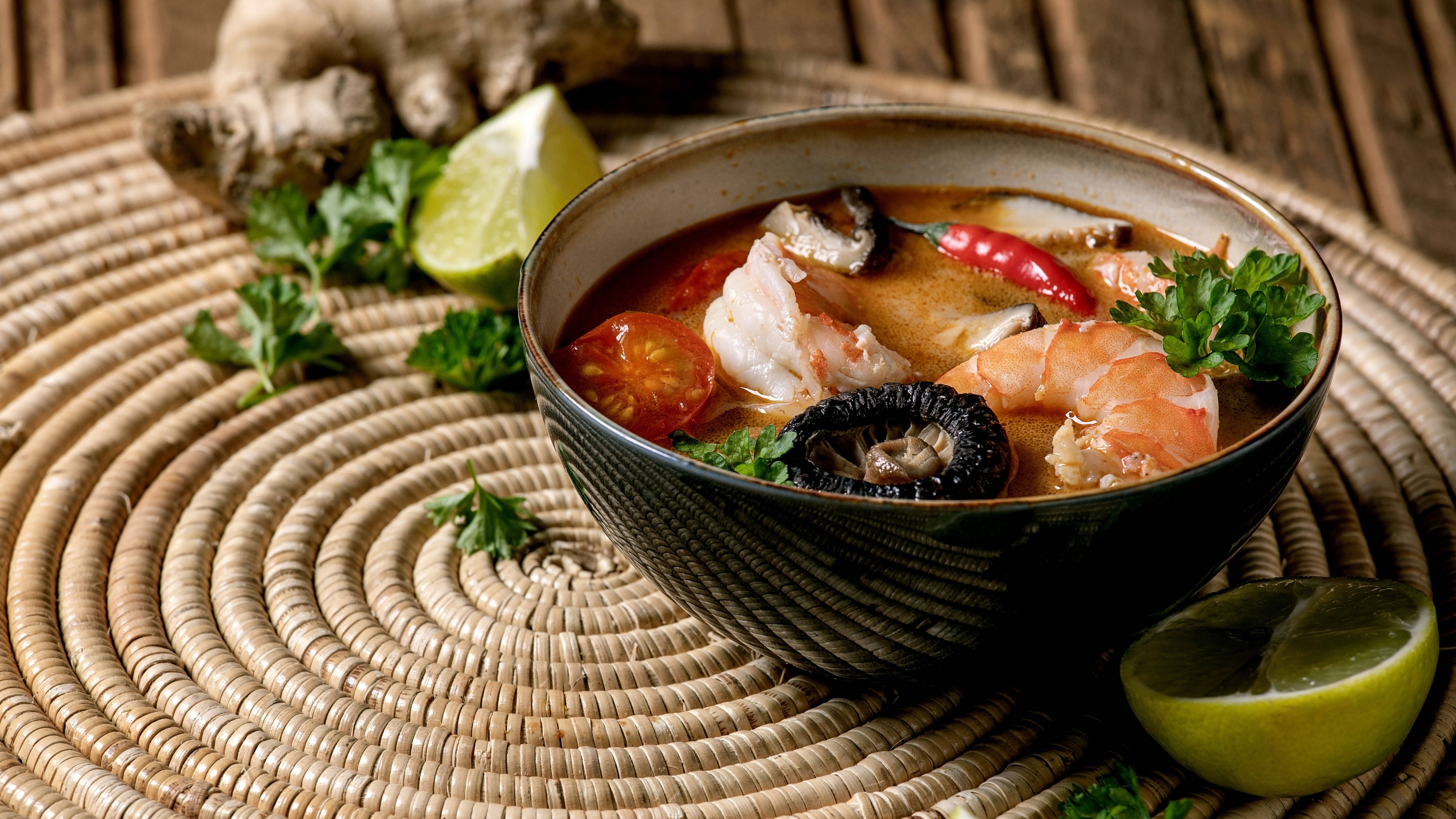 泰國經典酸辣湯Tom Yum 壁紙 — 一碗冒煙的辣冬陰功湯，有蝦、蘑菇和青檸，放在編織墊上，周圍環繞著新鮮香草和生薑。