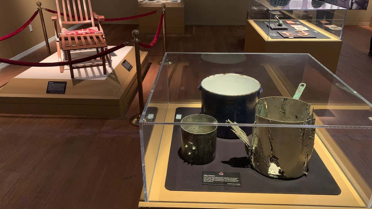 泰坦尼克號博物館內展出的珍貴文物 壁紙 — 磨損的金屬鍋和平底鍋陳列在博物館展覽的玻璃櫃中，展廳有木地板和用繩索隔開的展示區域。