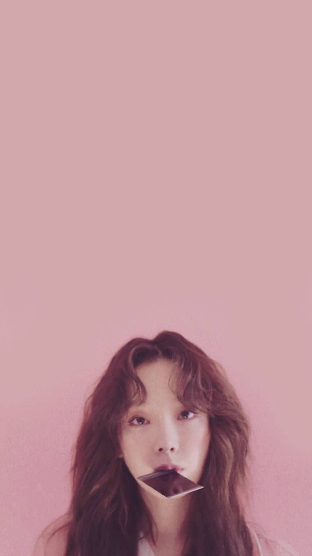 泰妍咬著寶麗來相片 壁紙 — 一位年輕女性，波浪形棕色頭髮，嘴裡含著一條黑朱古力棒，背景為純粉紅色。