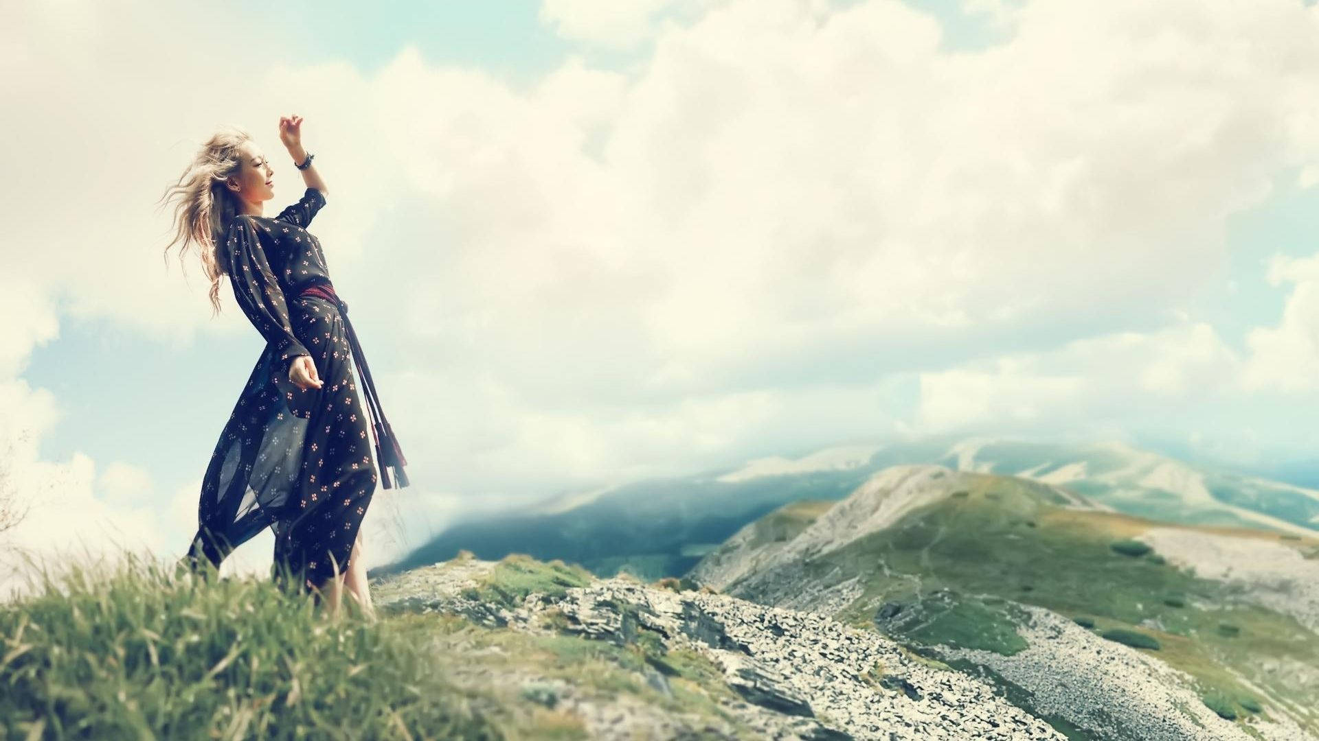 泰妍山頂風采 壁紙 — 一位身穿飄逸深色連衣裙的女性赤腳站在長滿青草的山脊上，向明亮的雲天舉起手。