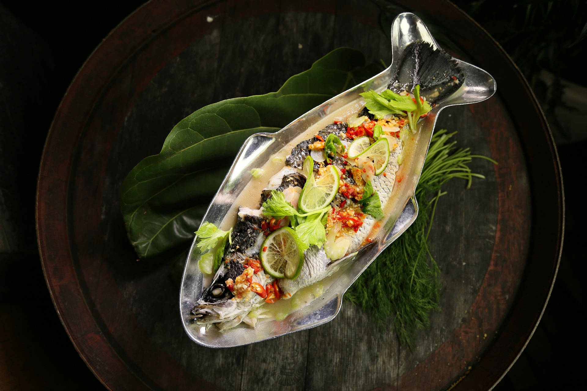 泰式巴拉米魚辣味料理 壁紙 — 一條完整嘅蒸魚，用青檸片、辣椒同香草裝飾，放喺魚形銀碟上，再擺喺深色木桶上面。