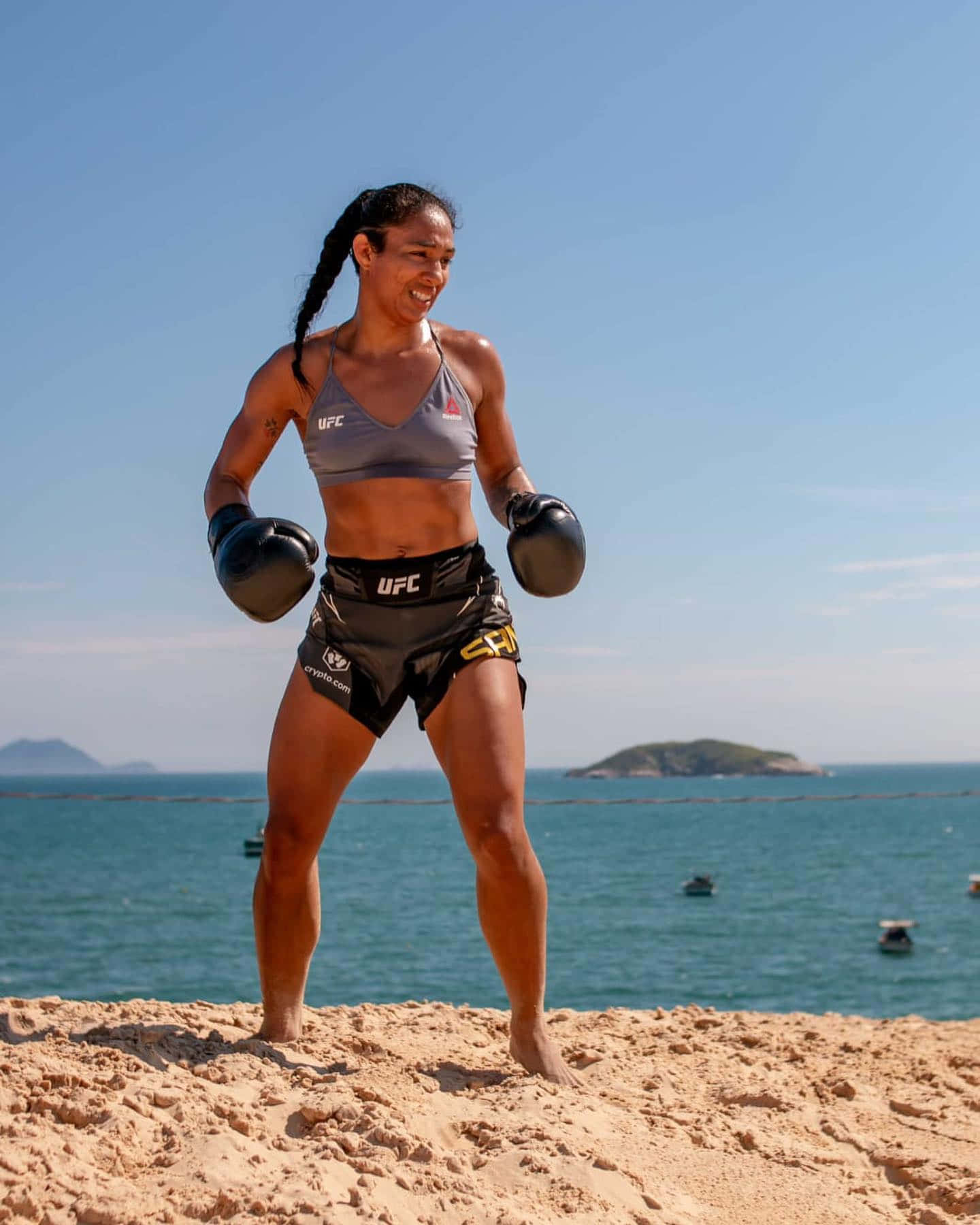 身穿UFC裝備的女性MMA拳手在陽光海灘上微笑，背景有海洋和島嶼。