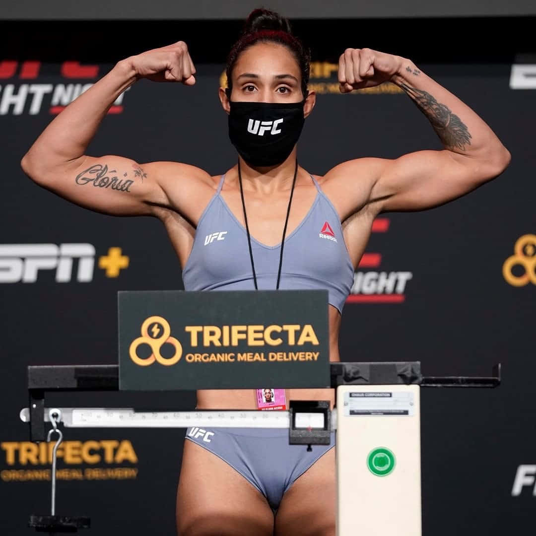 女UFC拳手在稱重時彎曲手臂，戴著口罩和灰色裝備。