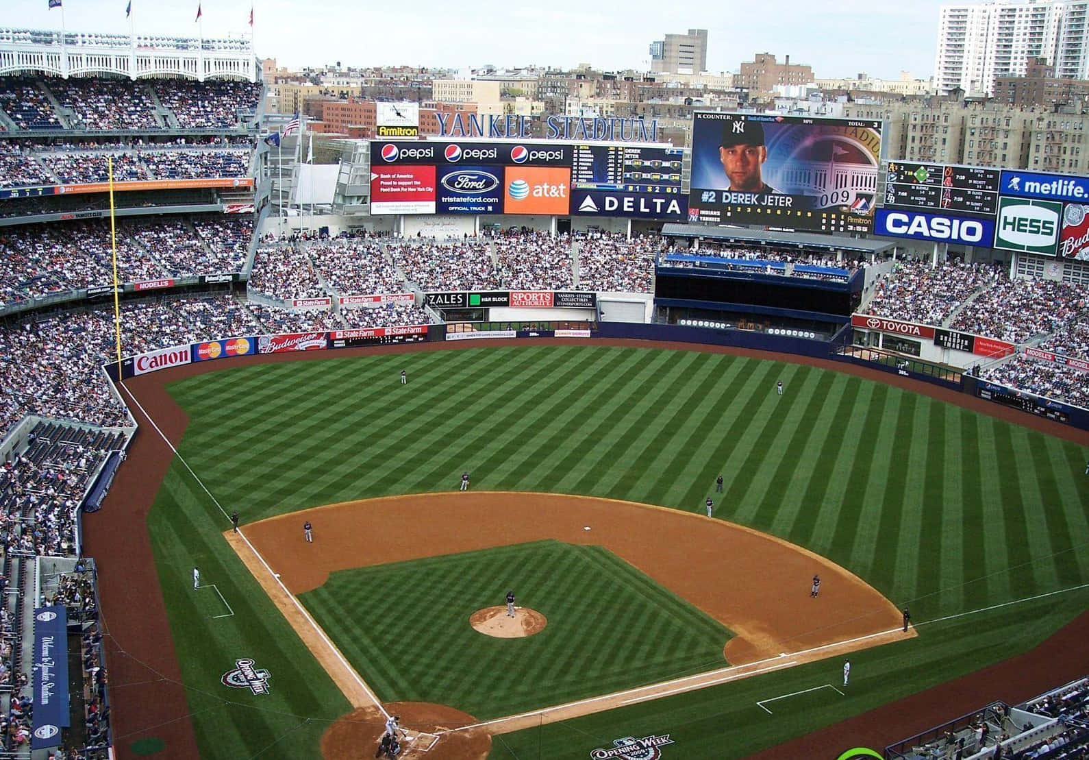 洋基球場棒球場景觀背景 壁紙 — 日間棒球比賽期間，洋基球場（Yankee Stadium）擠滿球迷，球員在條紋綠色場地上的鳥瞰圖。