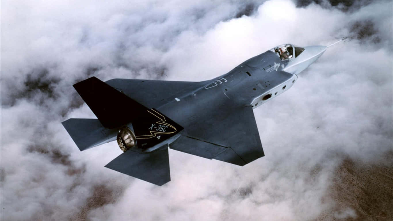 洛克希德·馬丁 X-35 飛行於晴空中的巨型客機背景（1366x768） 壁紙 — 一架流線型灰色X-35C戰鬥機在濃密的白雲上方翱翔，駕駛艙內可見飛行員。