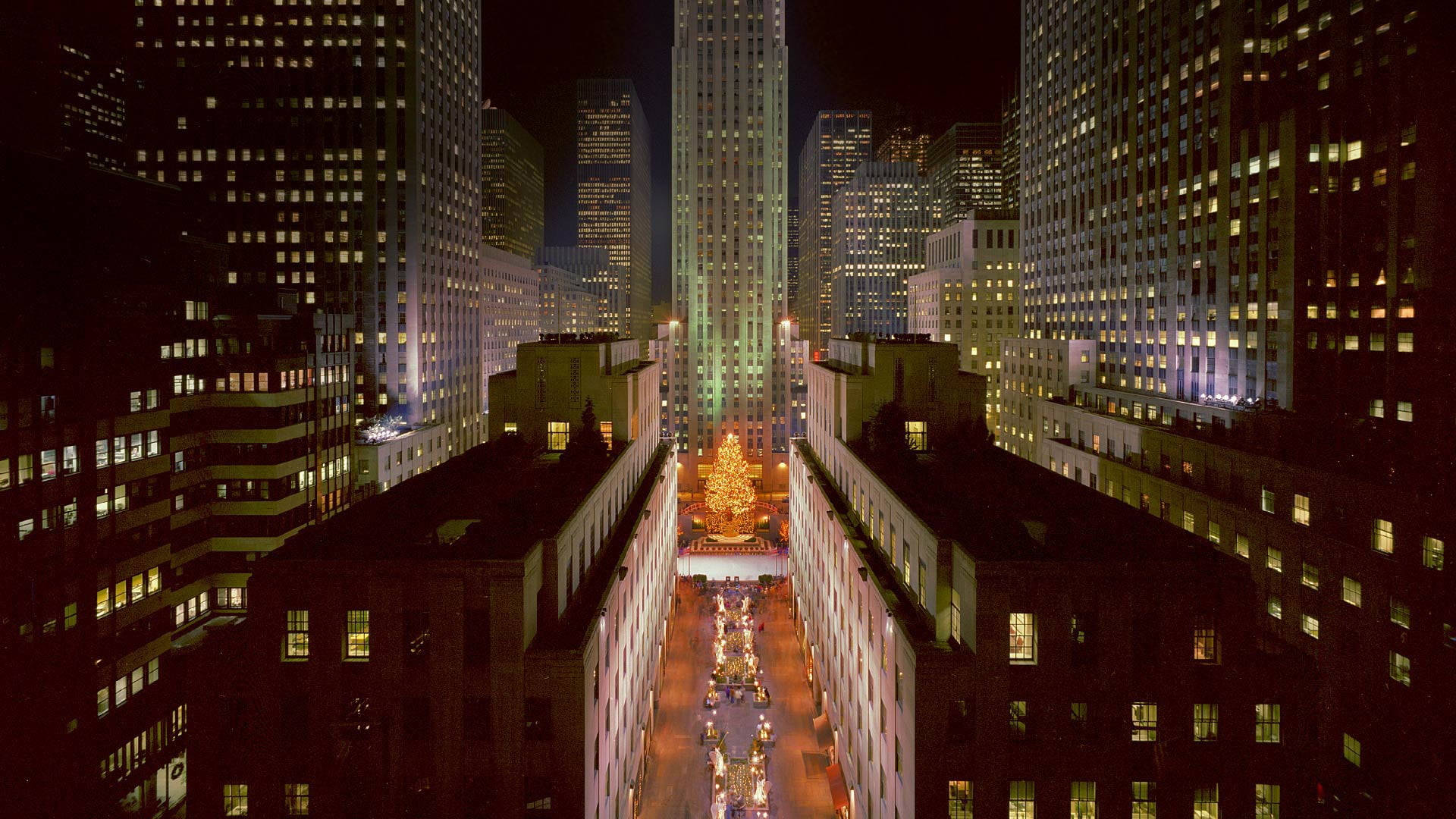 洛克菲勒中心建築夜景 壁紙 — 夜晚洛克菲勒中心的鳥瞰圖，閃耀的聖誕樹和燈火通明的摩天大樓環繞著熙攘的廣場。