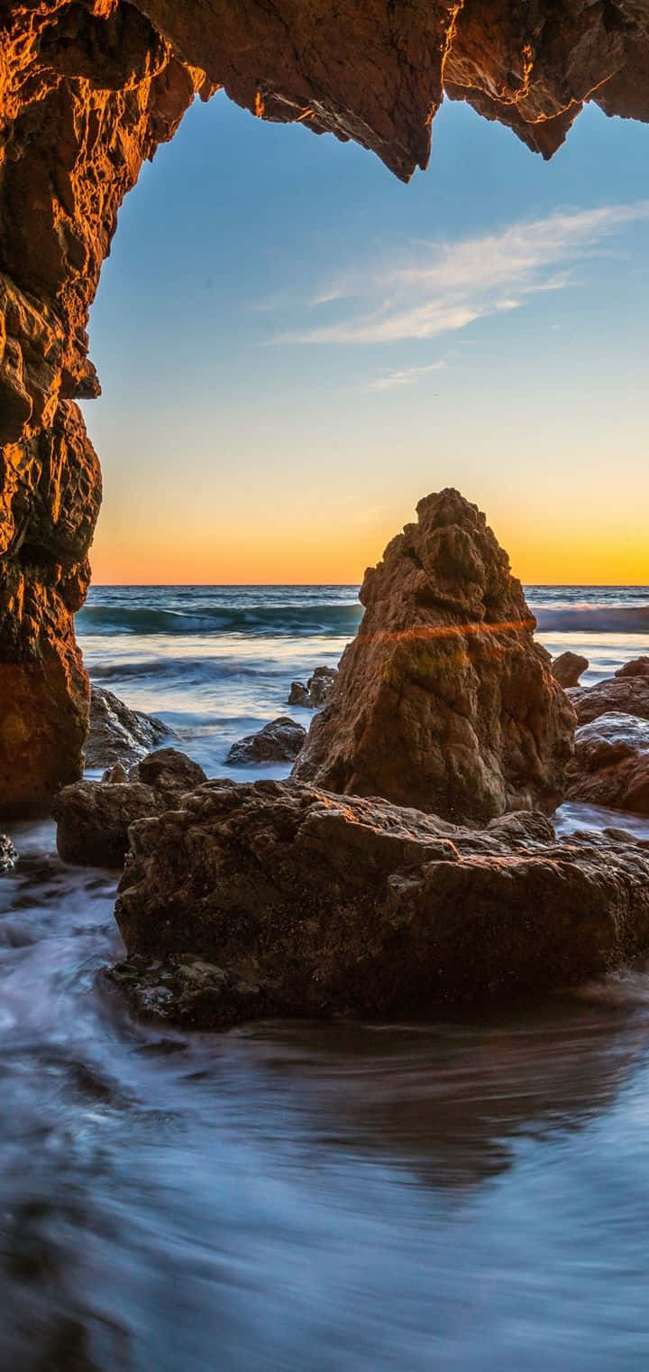 洞穴與夕陽天際線的安卓馬利布背景 壁紙 — 一個天然岩石拱門框住寧靜的海洋日落，金色光線反射在紋理豐富的懸崖和溫柔的海浪上。