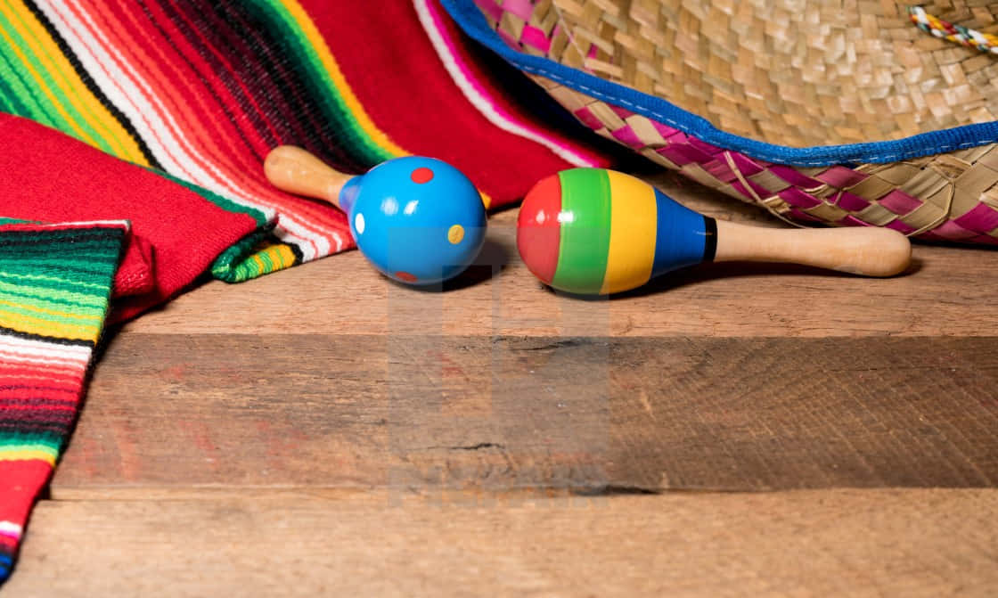 活力四射 Cinco de Mayo 節慶背景 壁紙 — 色彩繽紛的沙槌放在質樸的木質表面上，旁邊有一條彩色條紋毯子和一頂編織帽。