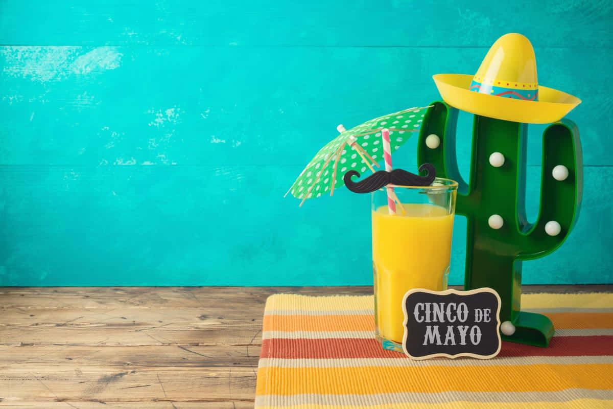 活力四射 Cinco de Mayo 節慶背景 壁紙 — 一杯節日氣氛濃厚的墨西哥五月五日節飲品，配有鬍子雨傘和仙人掌裝飾，放在條紋墊子上，背景是藍綠色木牆。
