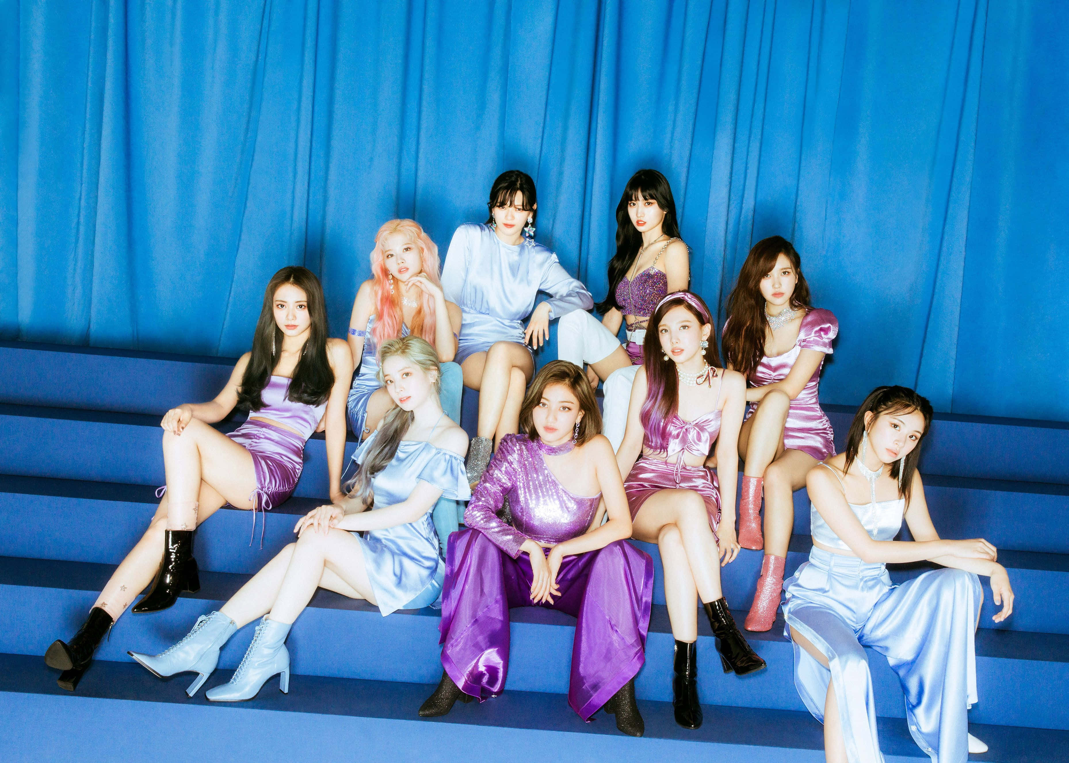 活力四射的 K-pop 組合演出 壁紙 — 七位身穿閃亮紫色和藍色服裝的女性在藍色台階上自信地擺姿勢，背景是相配的窗簾。