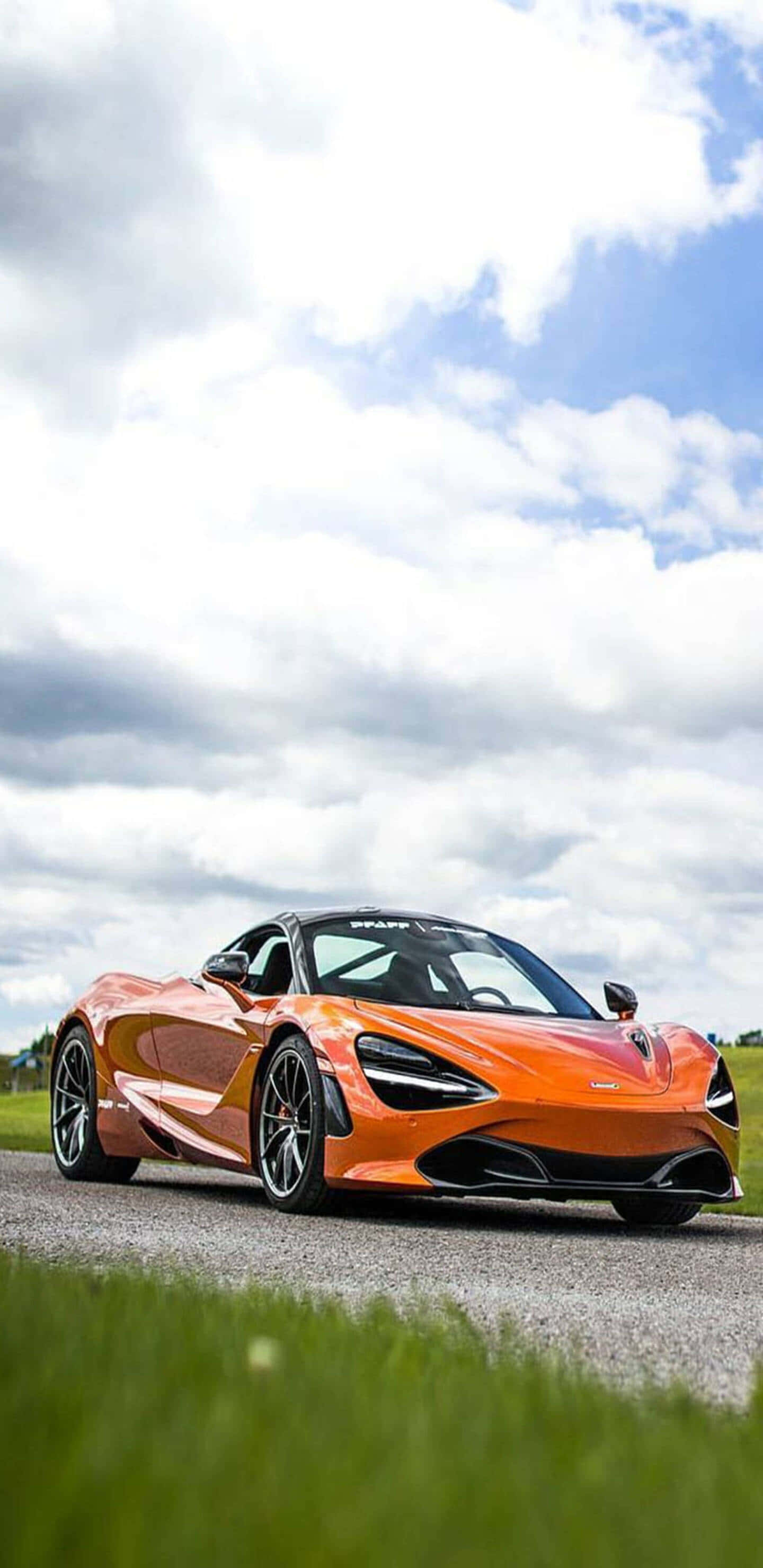 活力四射的麥拉倫720S於Pixel 3XL螢幕展示 壁紙 — 一輛充滿活力的橙色McLaren 720S跑車在部分多雲的天空下，在綠草如茵的碎石路上閃閃發光。