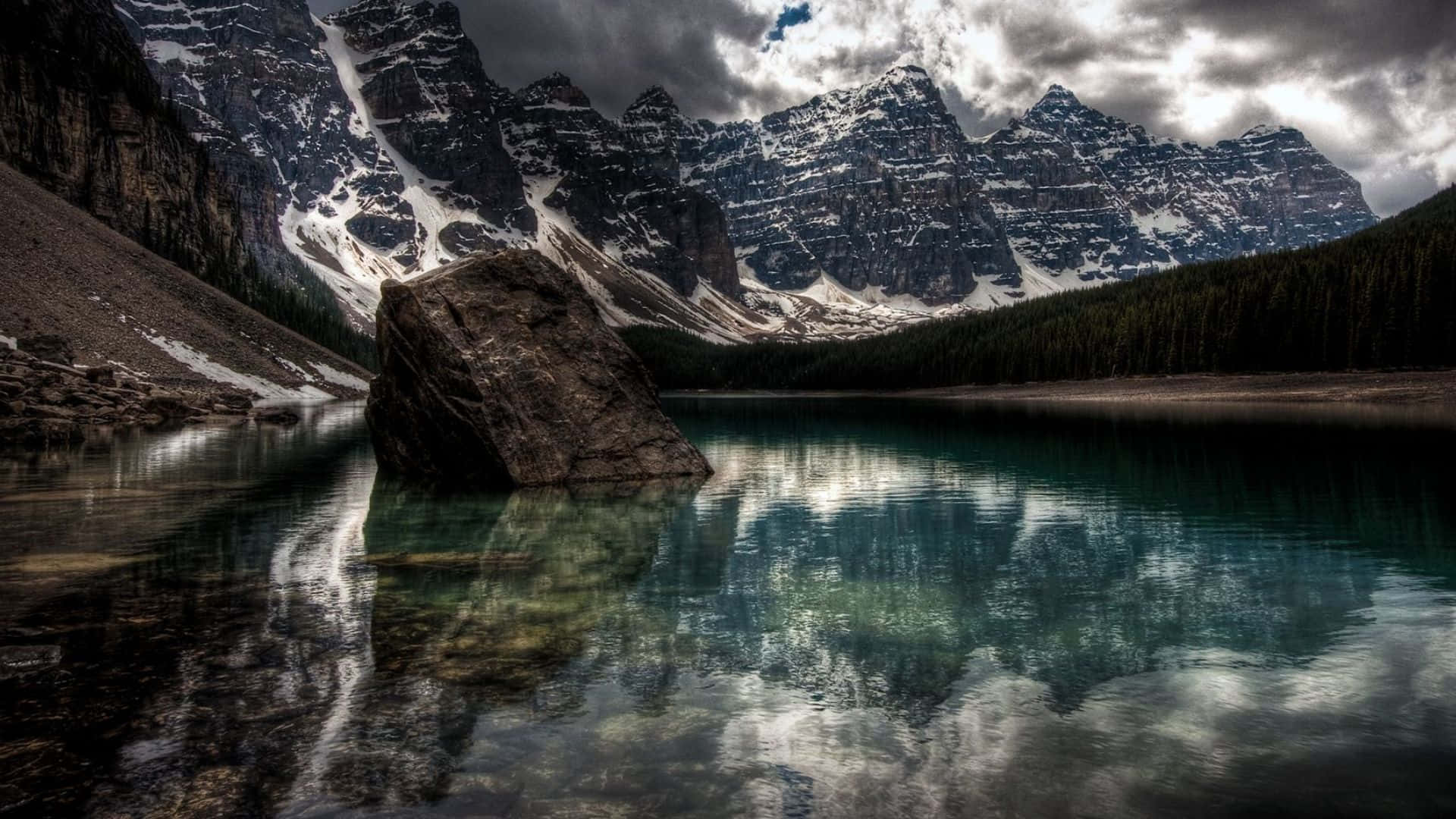 活力多彩全高清背景 壁紙 — 一塊孤獨的巨石坐落在清澈見底的湖泊中，倒映著白雪覆蓋的山峰，天空烏雲密布，氣氛戲劇化。
