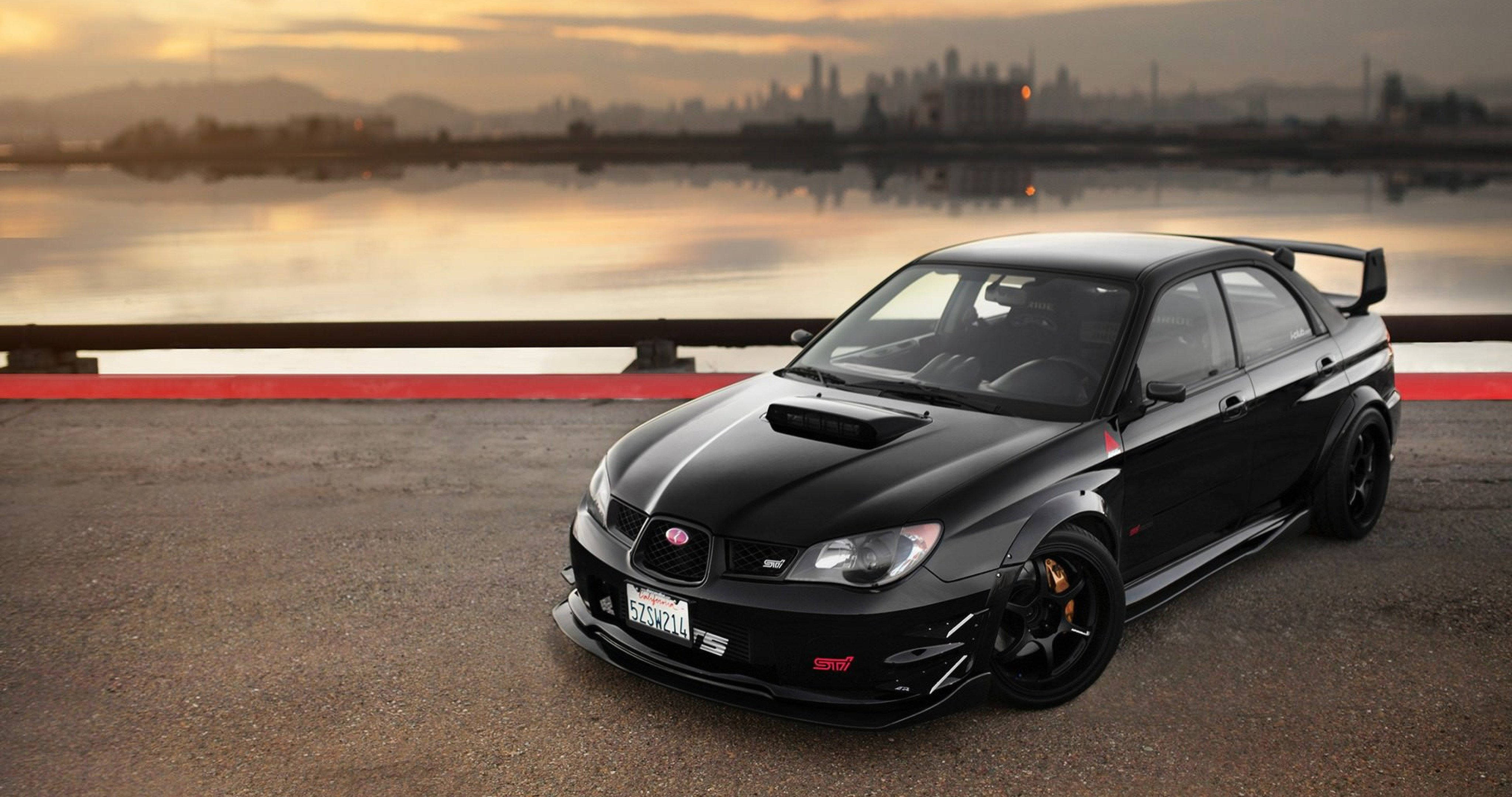 活力紅色斯巴魯Impreza徽章 壁紙 — 一架時尚的黑色Subaru WRX STI在日落時停泊在水邊，遠處有城市天際線。
